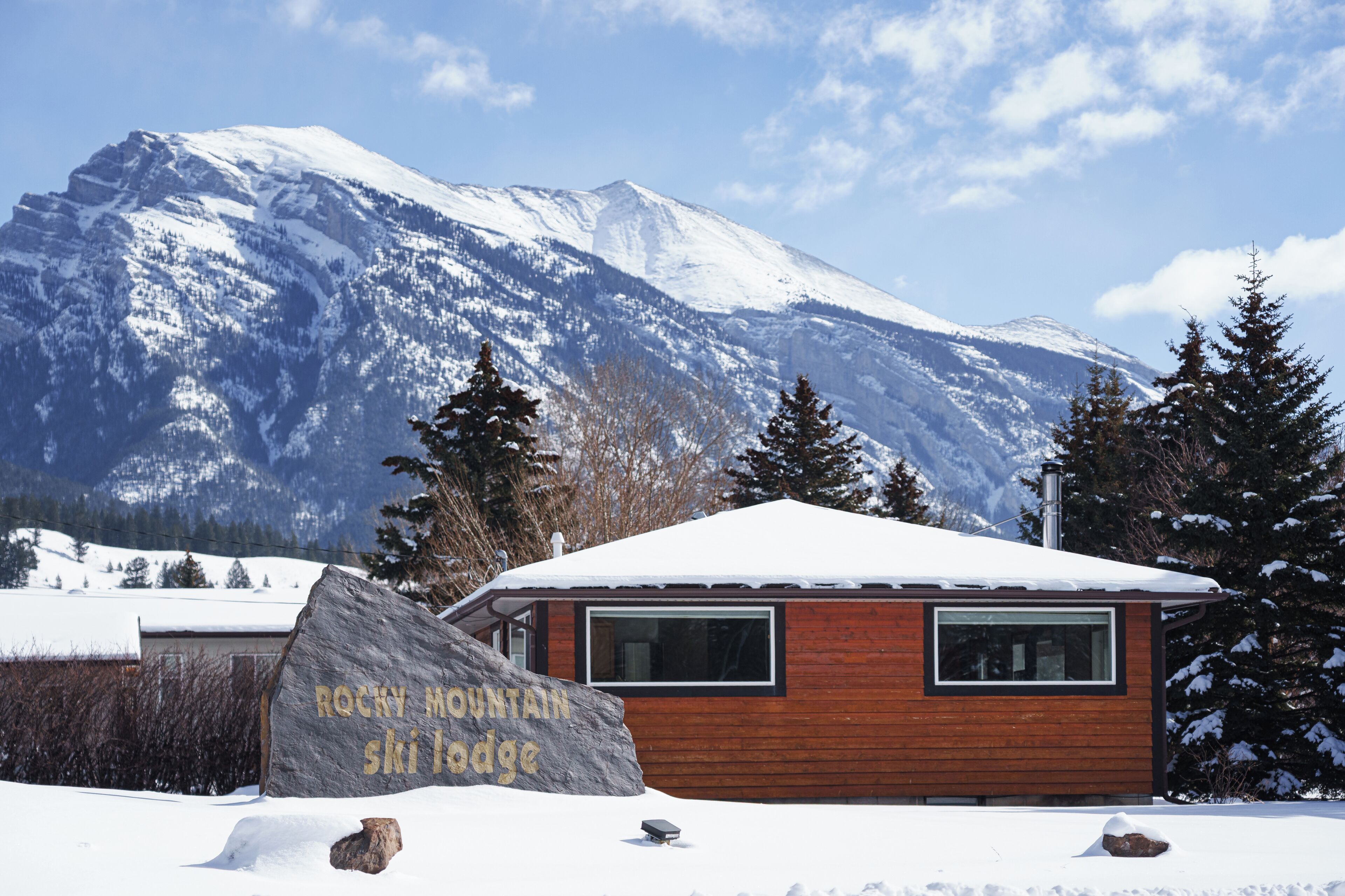 Een houten chalet met een met sneeuw bedekte dak, een grote rots waarop 'Rocky Mountain Ski Lodge' staat, en een berg op de achtergrond.