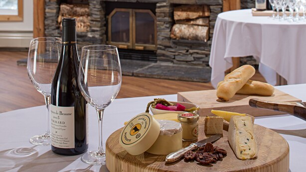 A room with a stone fireplace, wooden beams, a table with wine bottles, glasses, cheese, and bread. - Rocky Mountain Ski Lodge (Canmore)