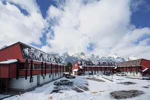 A hotel with red and white exterior walls, a parking area with snow, and a mountain backdrop.