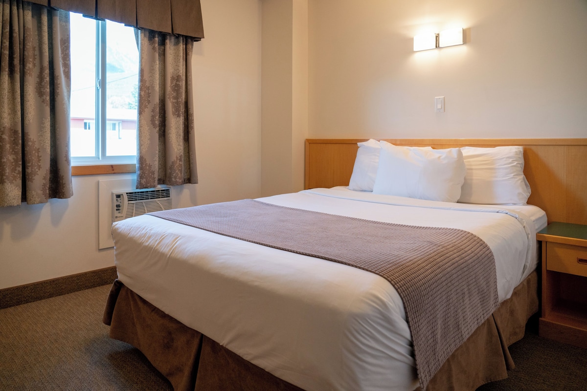 A neatly made bed with a wooden headboard, a bedside table, and a window with curtains.
