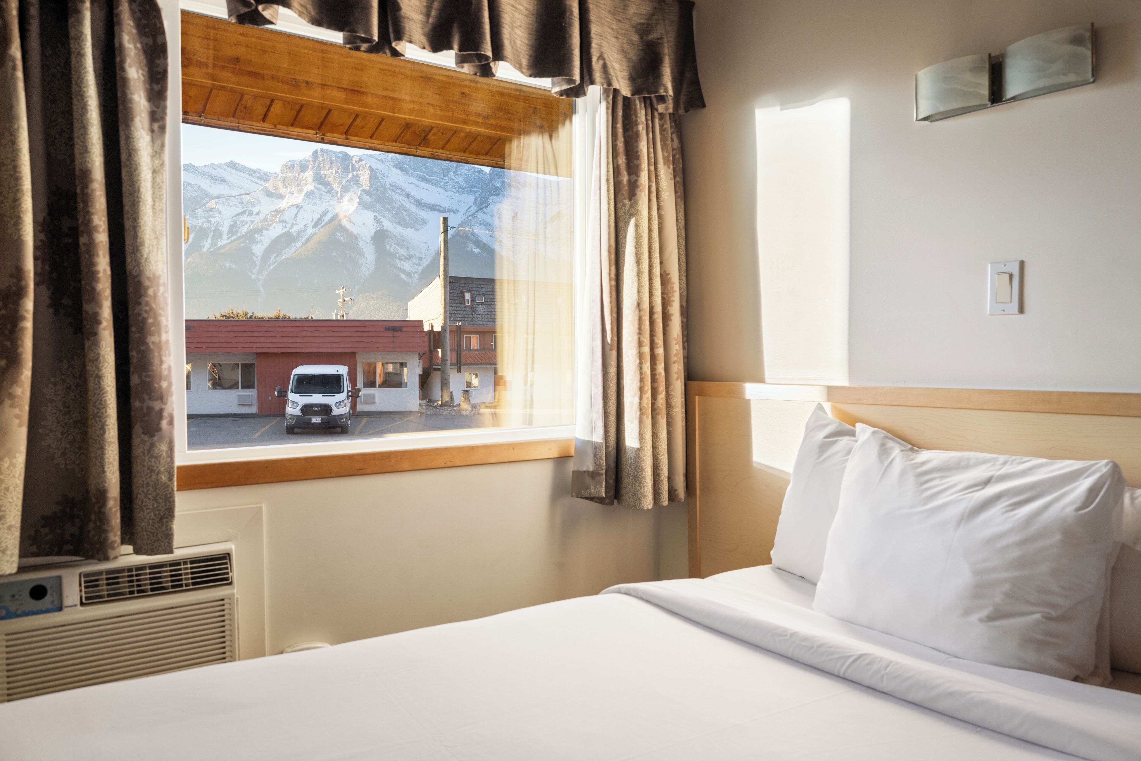 A hotel room with a bed, a window with curtains, a view of mountains, and a building outside.