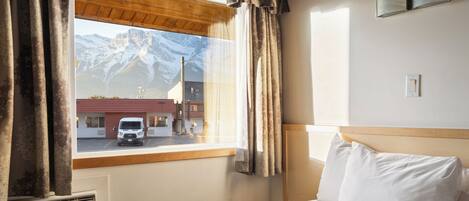 A hotel room with a bed, a window with curtains, a view of mountains, and a building outside.