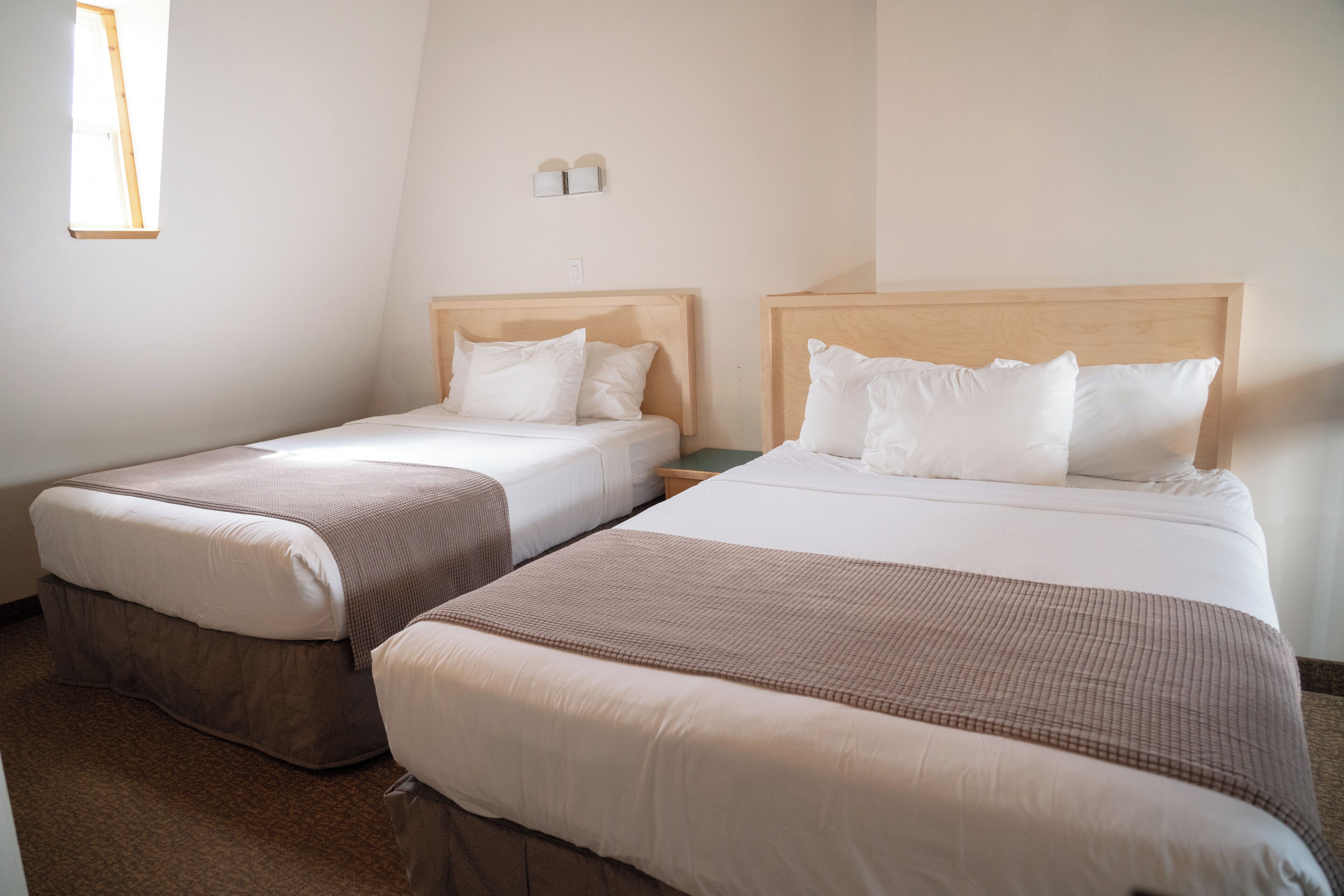 Dos camas individuales con cabeceros de madera, ropa de cama blanca y una ventana pequeña a la izquierda.
