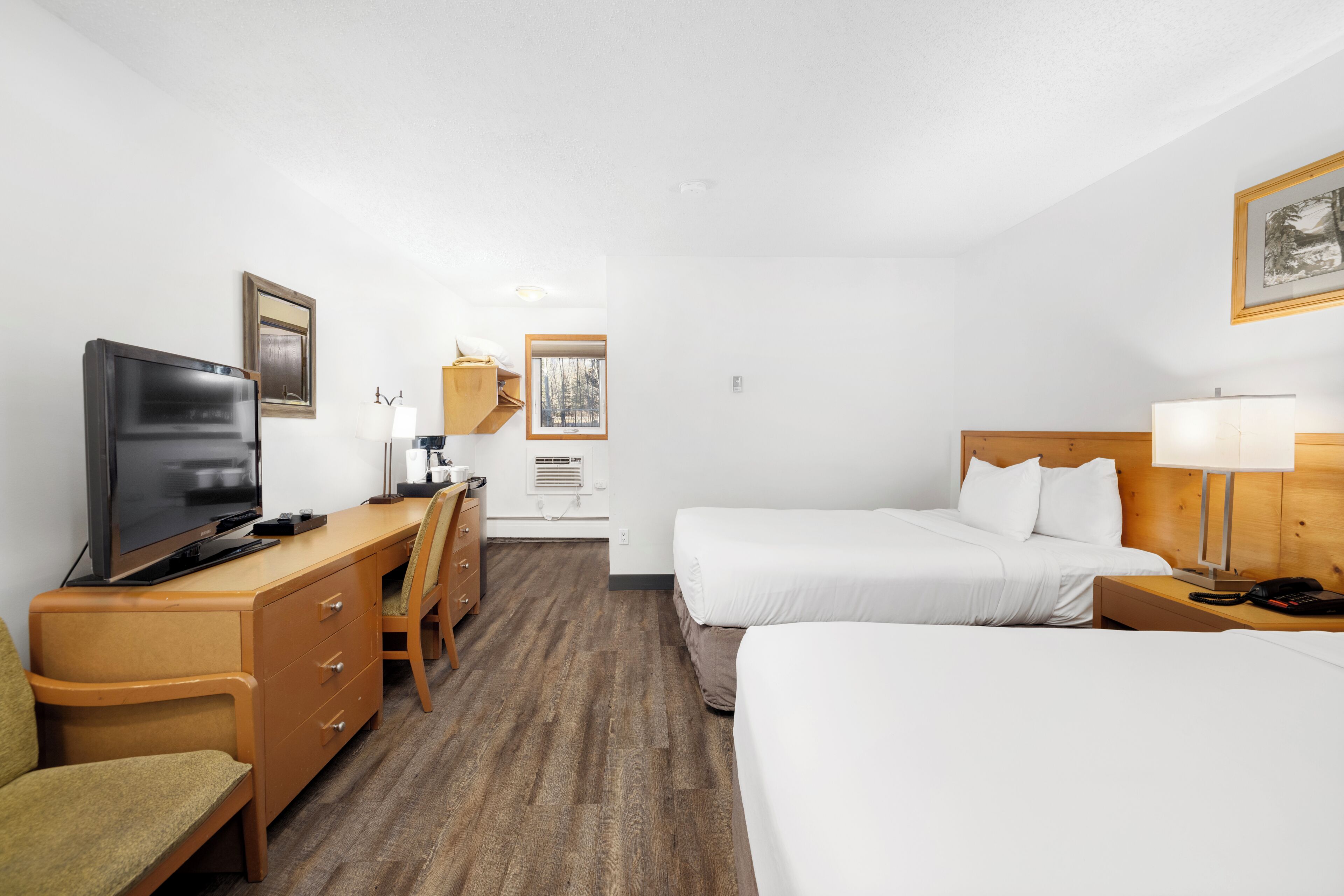 A hotel room with a bed, a desk with a television, a chair, and a bedside table with a lamp.
