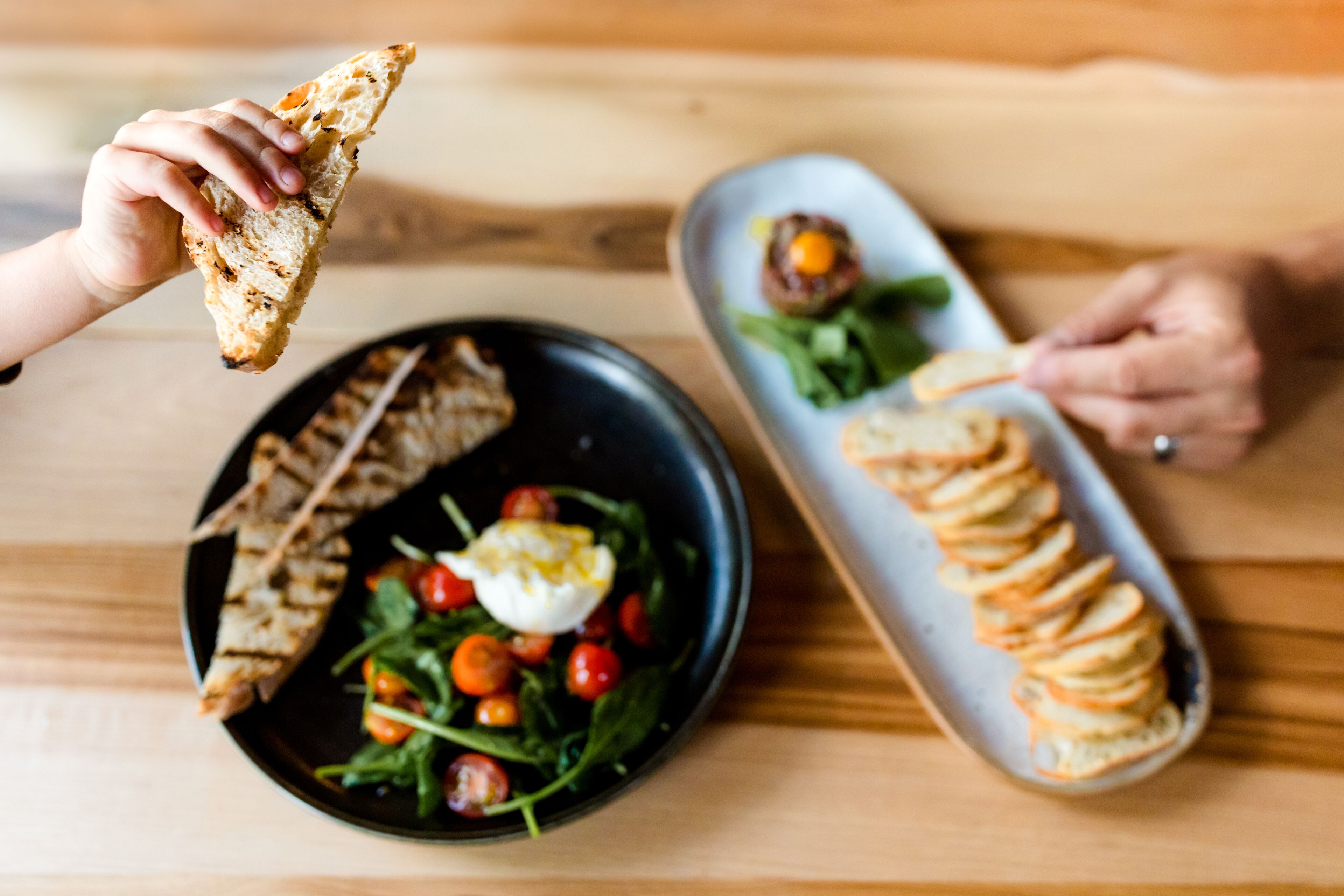 A dining table with a black plate containing a salad with cherry tomatoes and a poached egg, and a white plate with a stack of bread slices and a small dish of sauce.