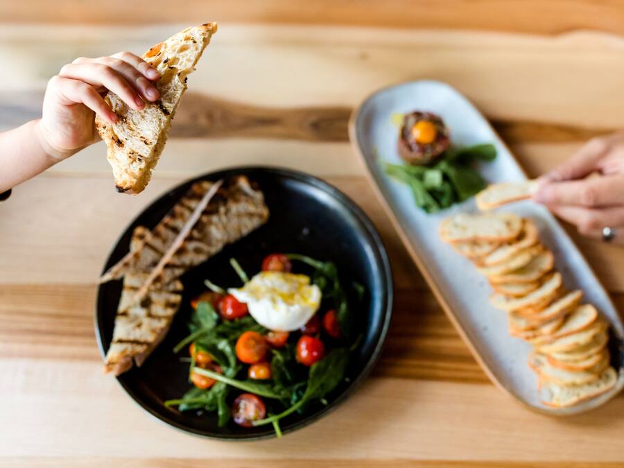 A dining table with a black plate containing a salad with cherry tomatoes and a poached egg, and a white plate with a stack of bread slices and a small dish of sauce.