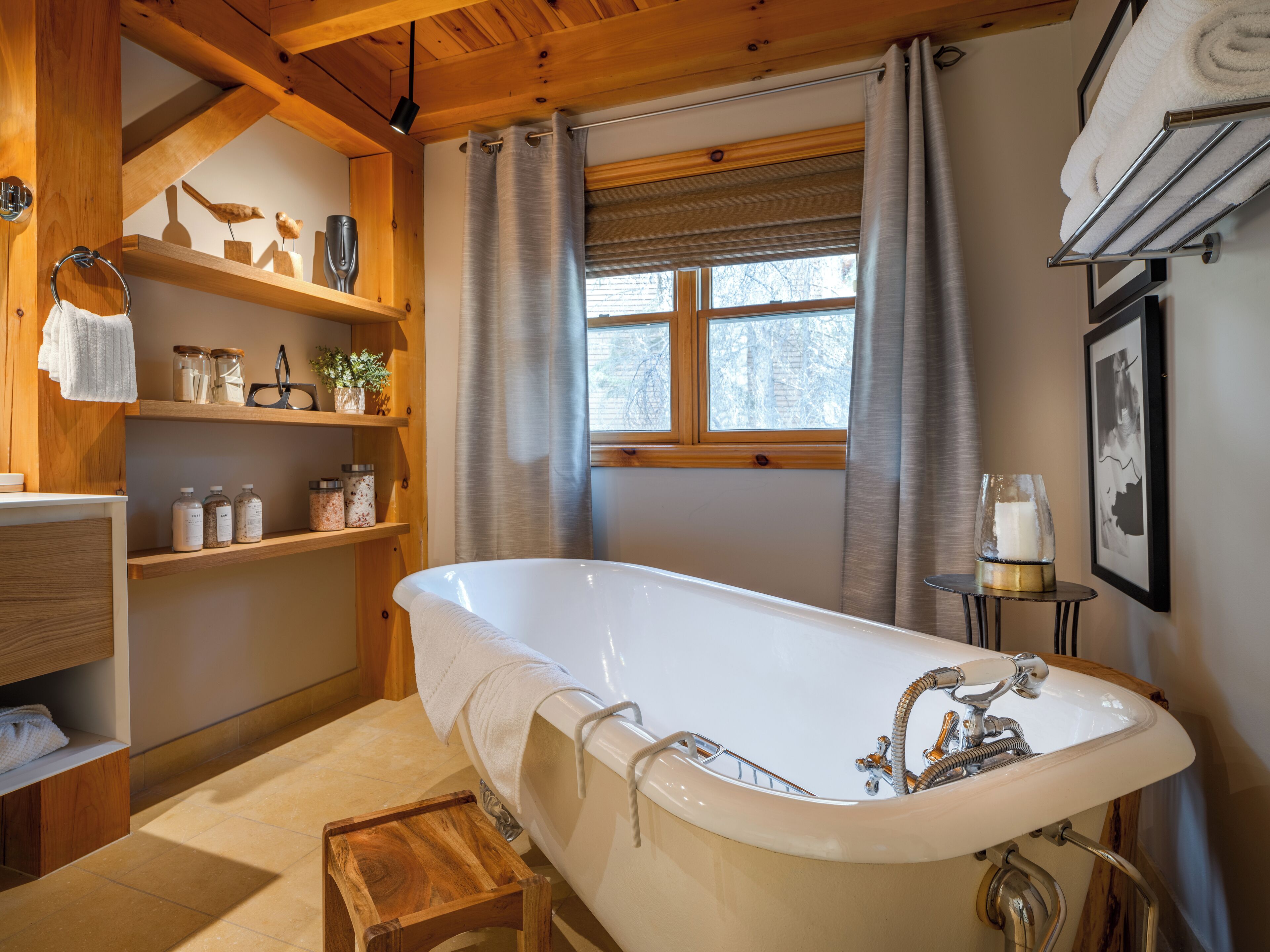 A bathroom with a freestanding bathtub, wooden shelves with jars, a window with curtains, and a framed picture on the wall.