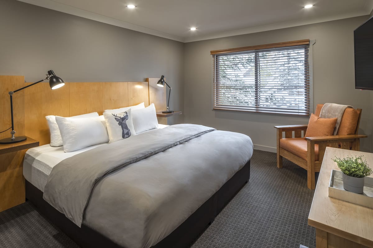 A modern hotel room with a large bed, a wooden headboard, a window with blinds, a chair, and a desk with a plant.