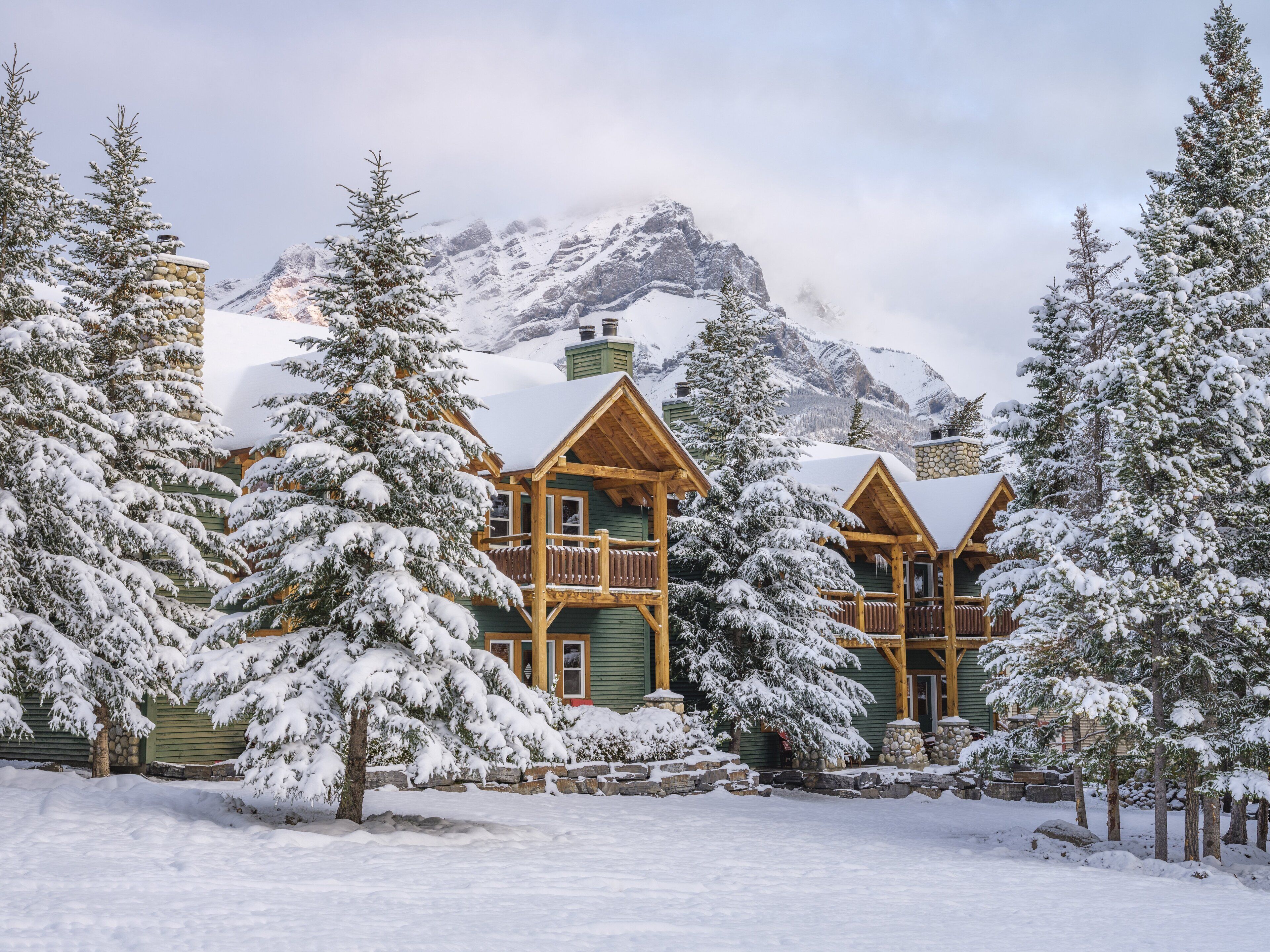 Une cabane en rondins dotée d’un balcon, entourée d’arbres couverts de neige et avec des montagnes en arrière-plan.