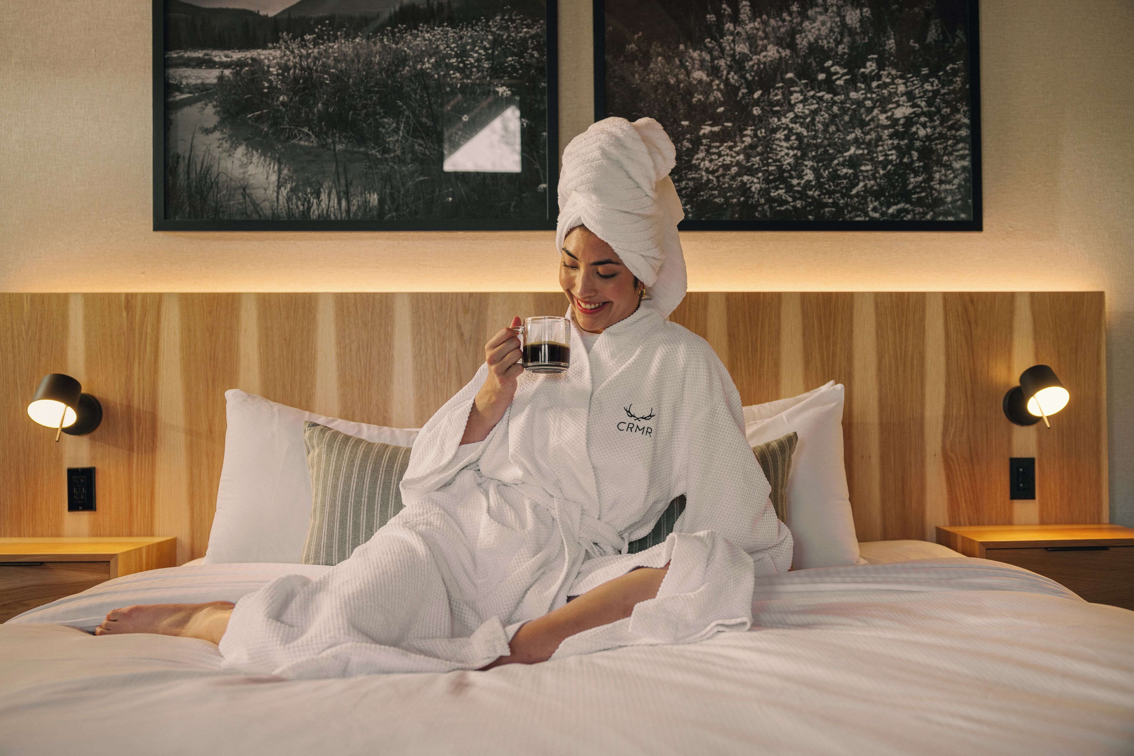 Eine Person in einem weißen Bademantel sitzt auf einem Bett mit einem Holz-Kopfschild und hält eine Tasse. Zwei Nachttischlampen und gerahmte Bilder sind an der Wand zu sehen.