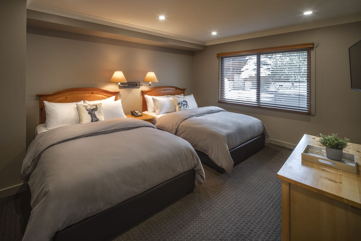 A bedroom with two beds, a nightstand with a lamp, a wooden desk, and a window with blinds.