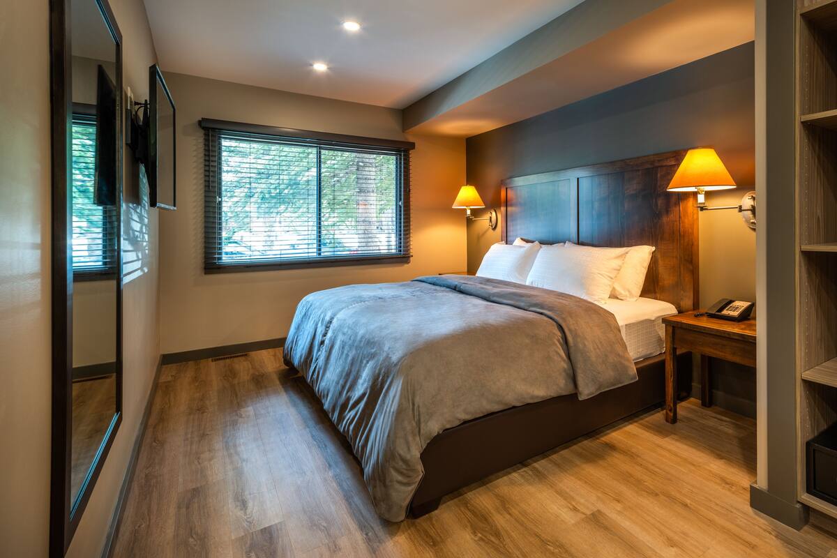 A modern bedroom with a wooden floor, a bed with a grey blanket, two bedside lamps, and a nightstand with a phone.