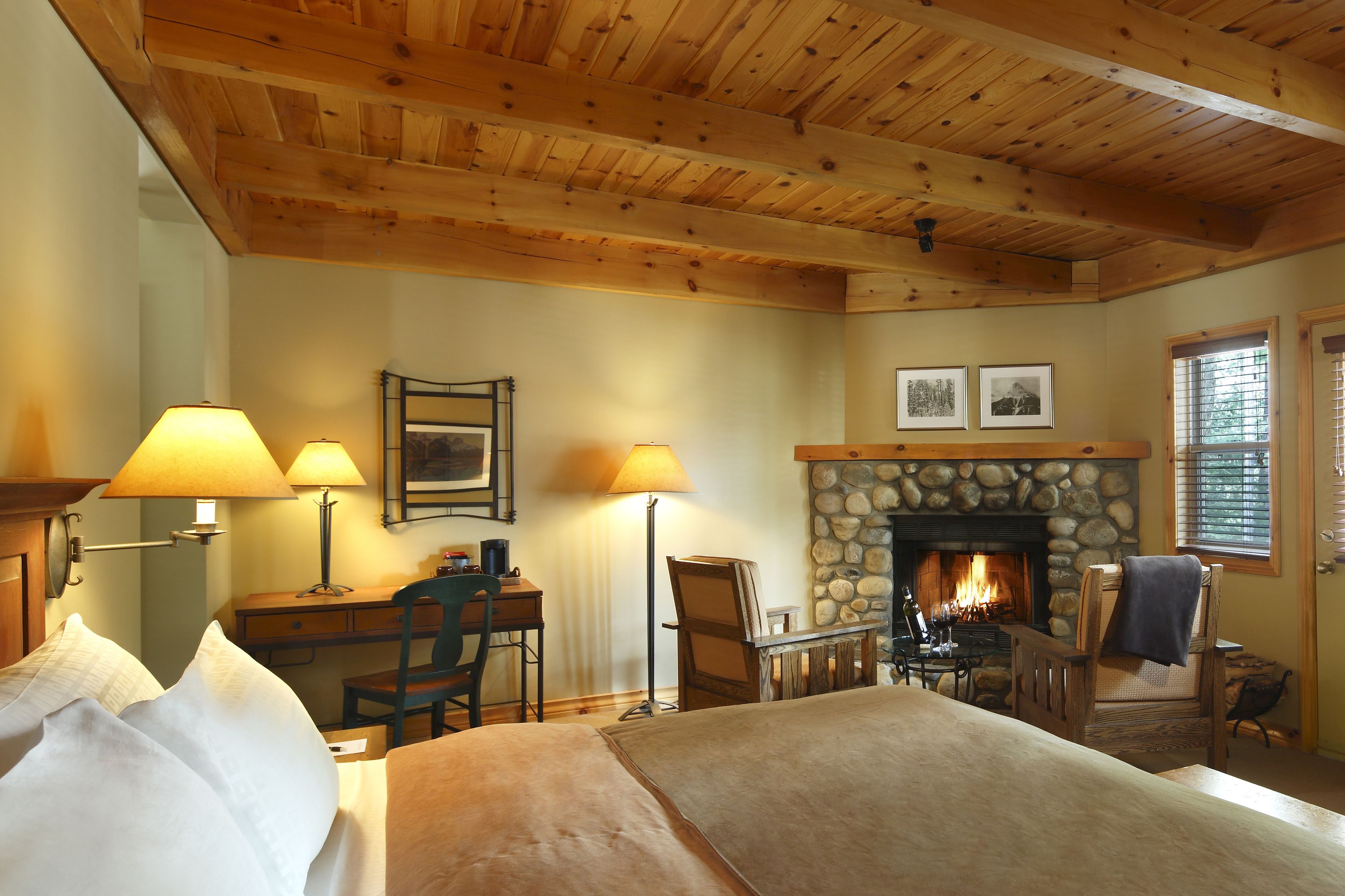 A cozy room with a bed, desk, chairs, a fireplace, and lamps.
