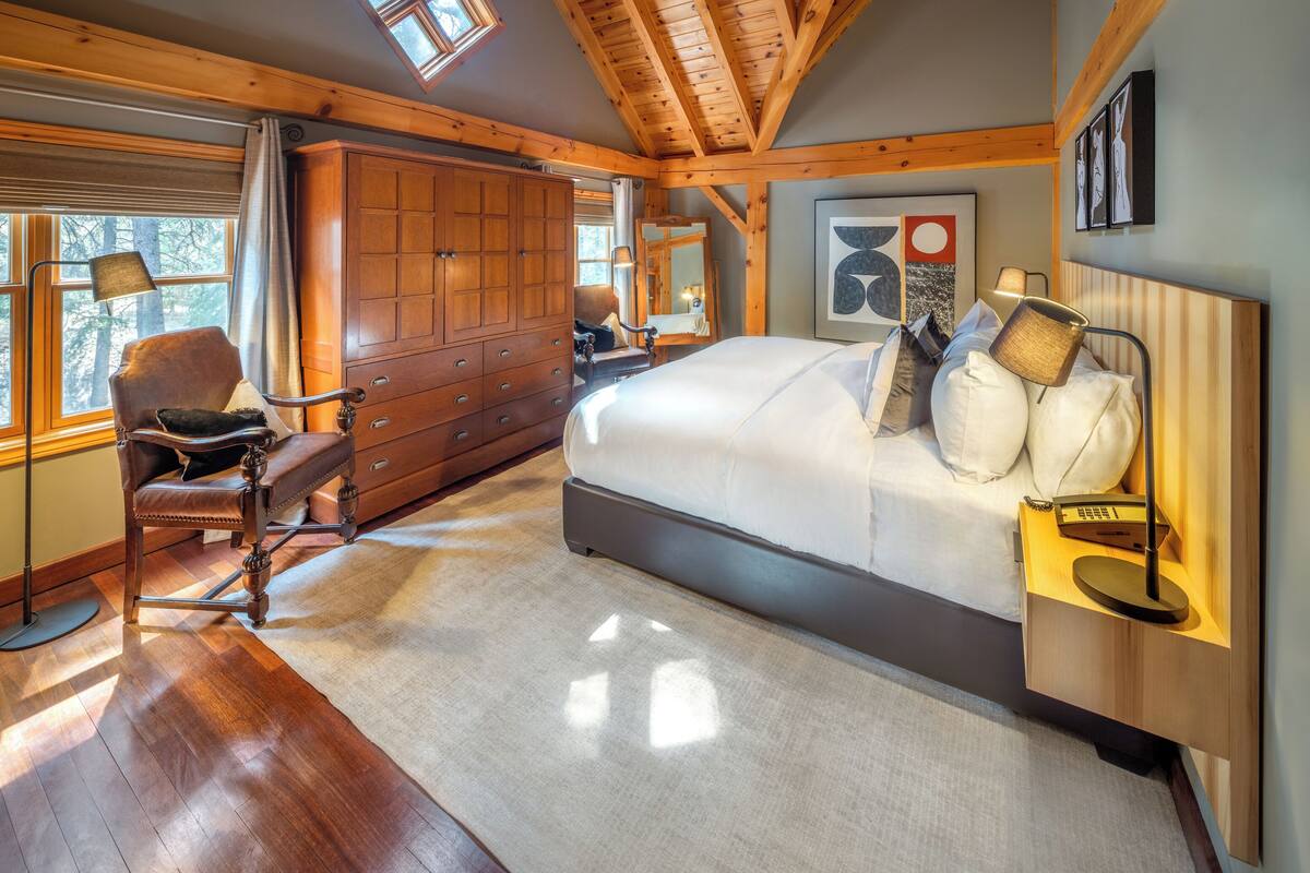 A spacious bedroom with a large bed, wooden ceiling beams, a window with curtains, and a nightstand with a lamp.