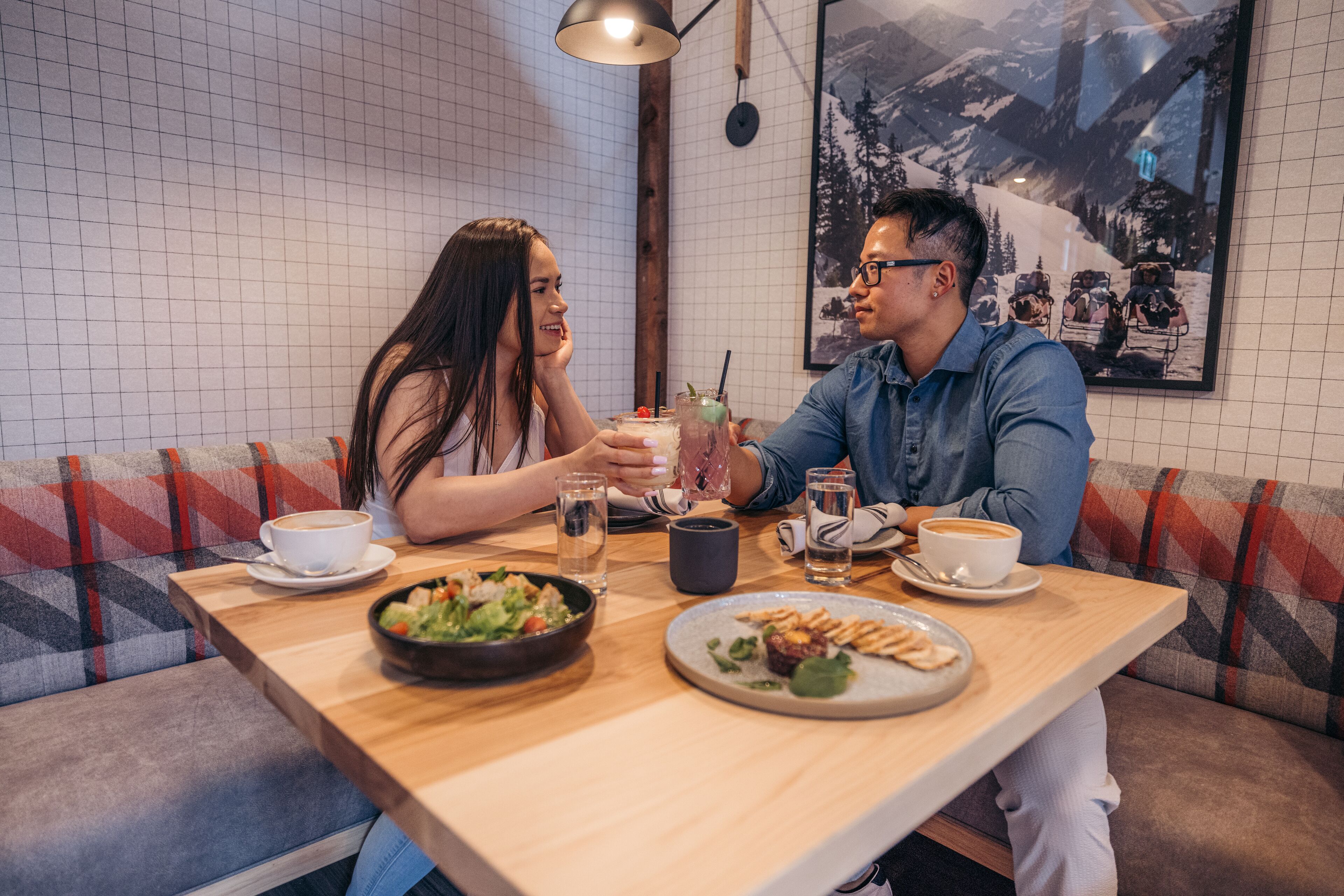 Una mesa de madera con dos personas sentadas, un tazón de ensalada, un plato de comida y tazas de café.