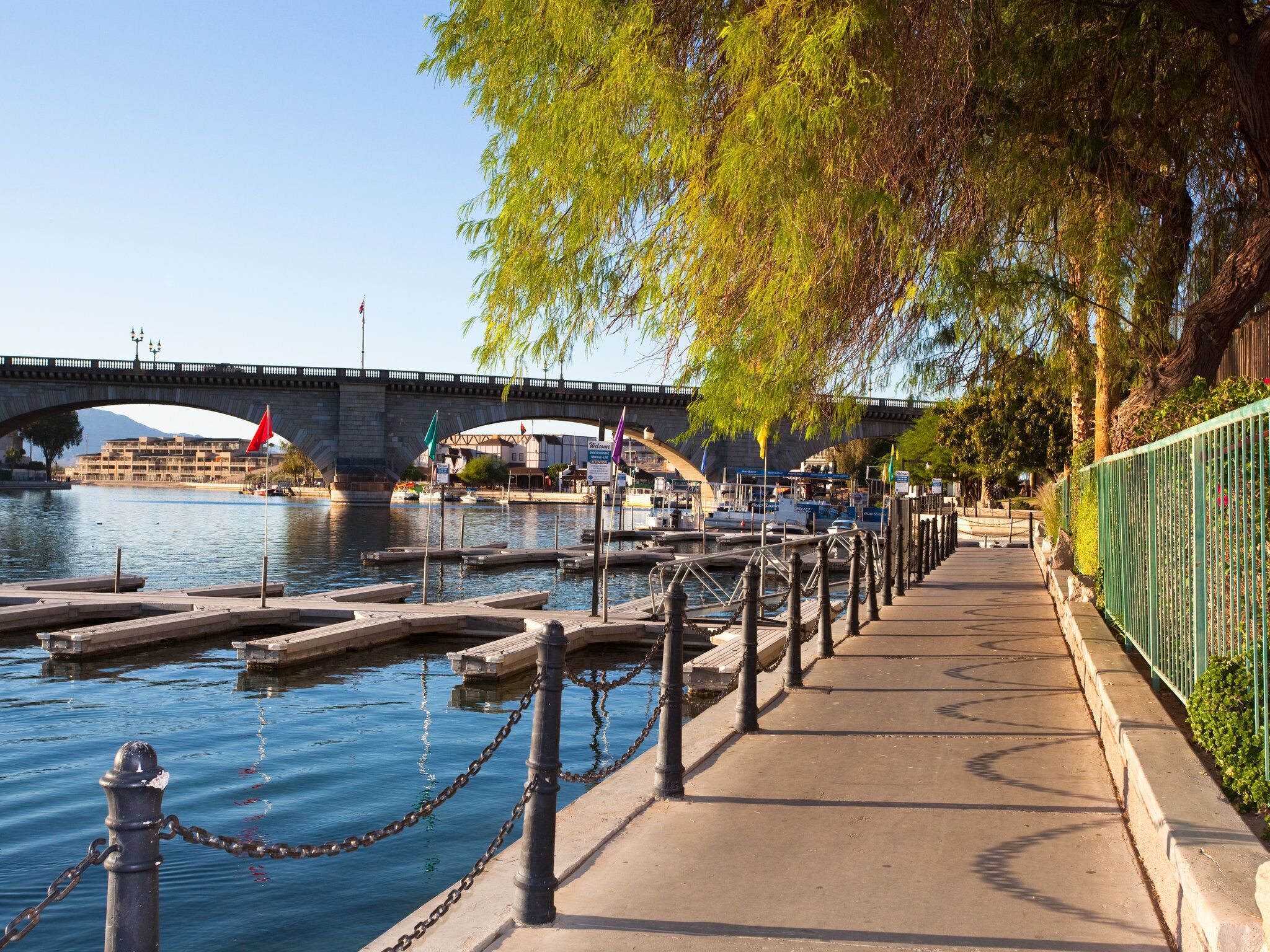 A riverside walkway with a stone bridge, boats docked along the river, and a green fence on the right.