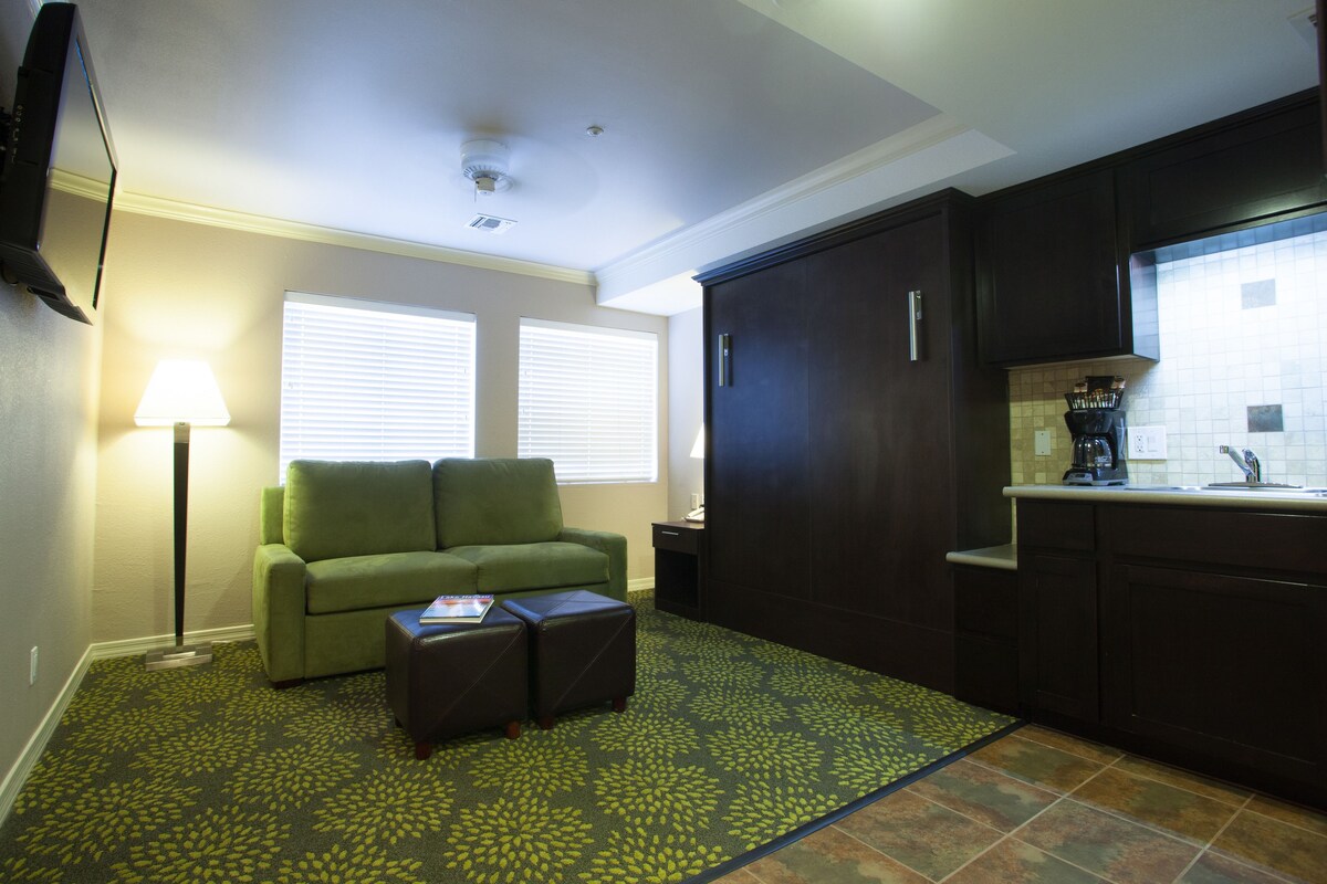 A compact living space with a sofa, ottoman, and a kitchen area with dark cabinets and a tiled backsplash.