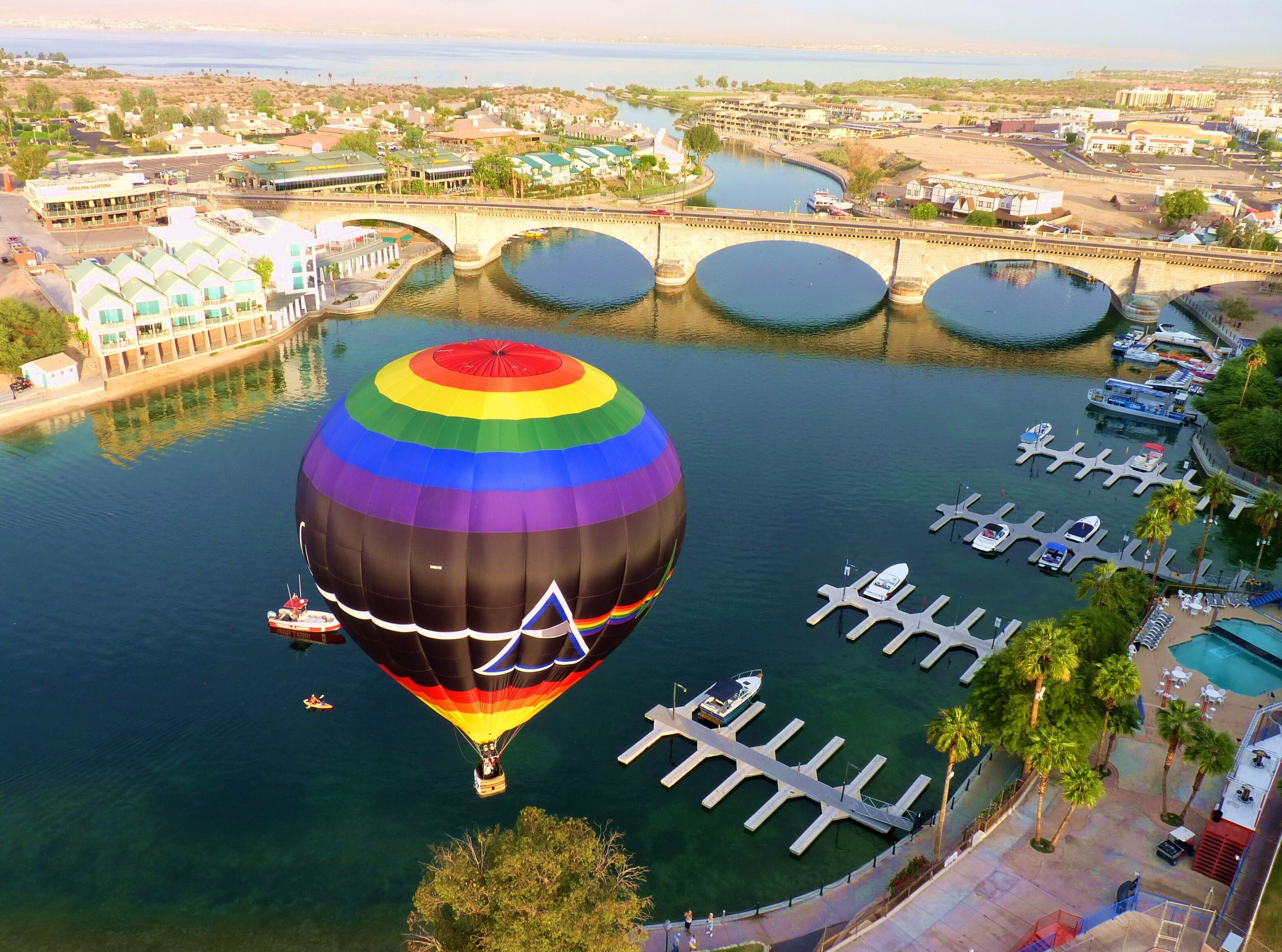 A hot air balloon over a marina with boats docked, a bridge, and buildings in the background.
