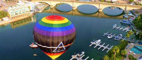 A hot air balloon over a marina with boats docked, a bridge, and buildings in the background.