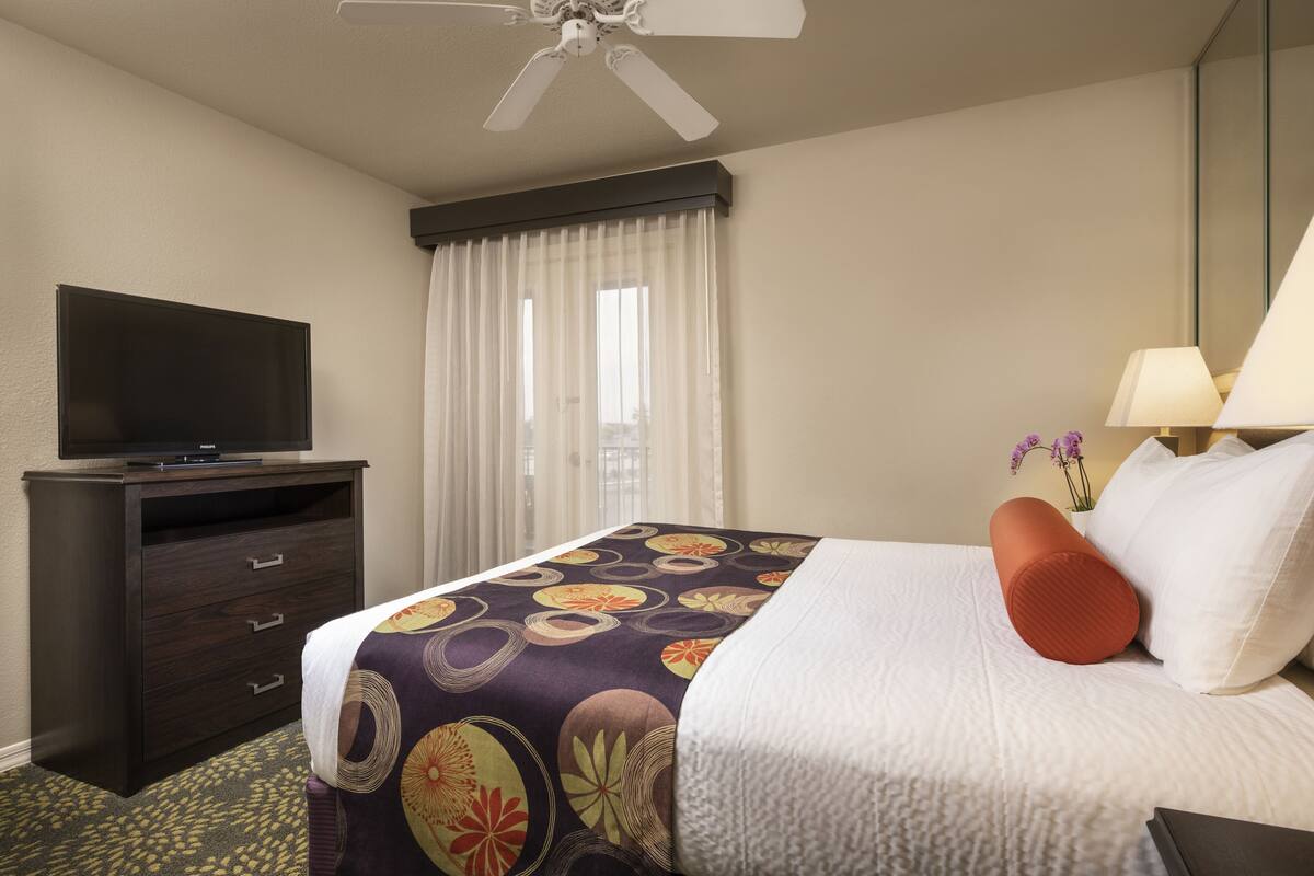 Habitación de hotel con cama, televisión en una cómoda, ventilador de techo y ventana con cortinas.