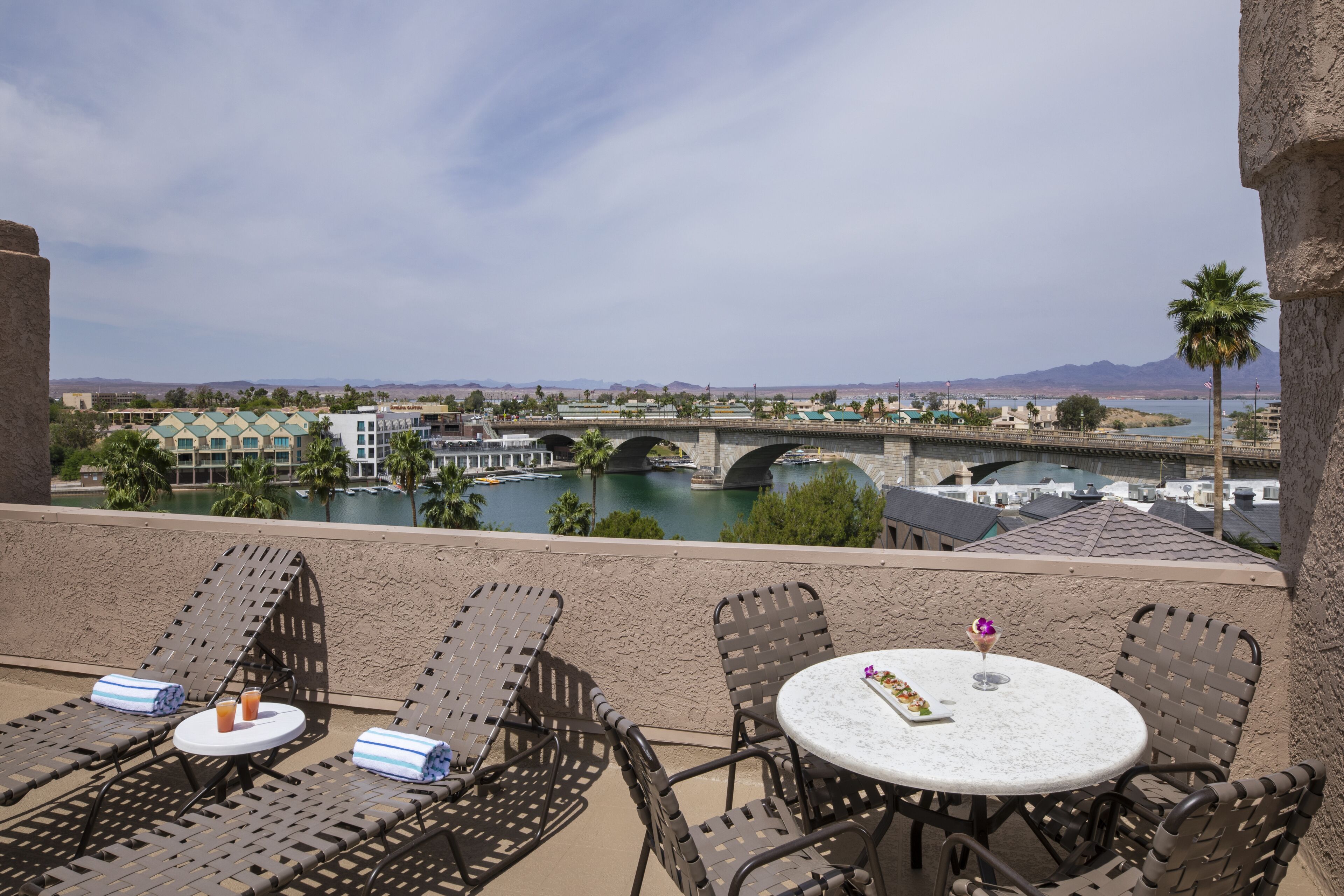 Een balkon met lounge fauteuils en een tafel, met uitzicht op een rivier met een brug en gebouwen op de achtergrond.