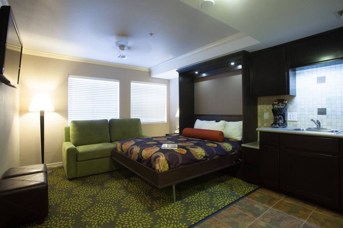 Una habitación de hotel compacta con cama, un sillón verde, una pequeña mesa, kitchenette y ventana con persianas.