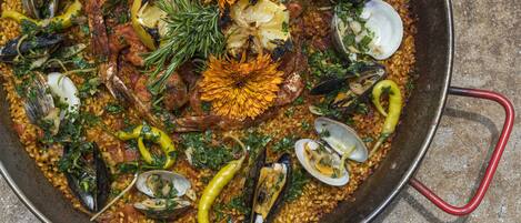 Une grande poêle ronde en métal, remplie de fruits de mer et de légumes, probablement une paella ou un plat similaire, avec une poignée sur le côté.