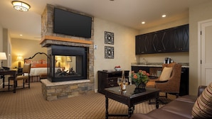 A living room with a fireplace, a television mounted above it, and a kitchen area with dark cabinets. - Casa Munras Garden Hotel & Spa (Monterey)