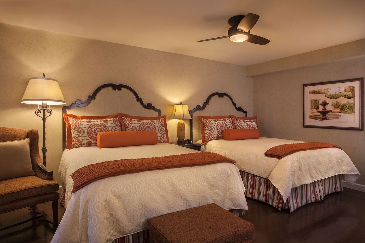 A hotel room with two beds, a ceiling fan, a lamp, a chair, and a framed picture on the wall.