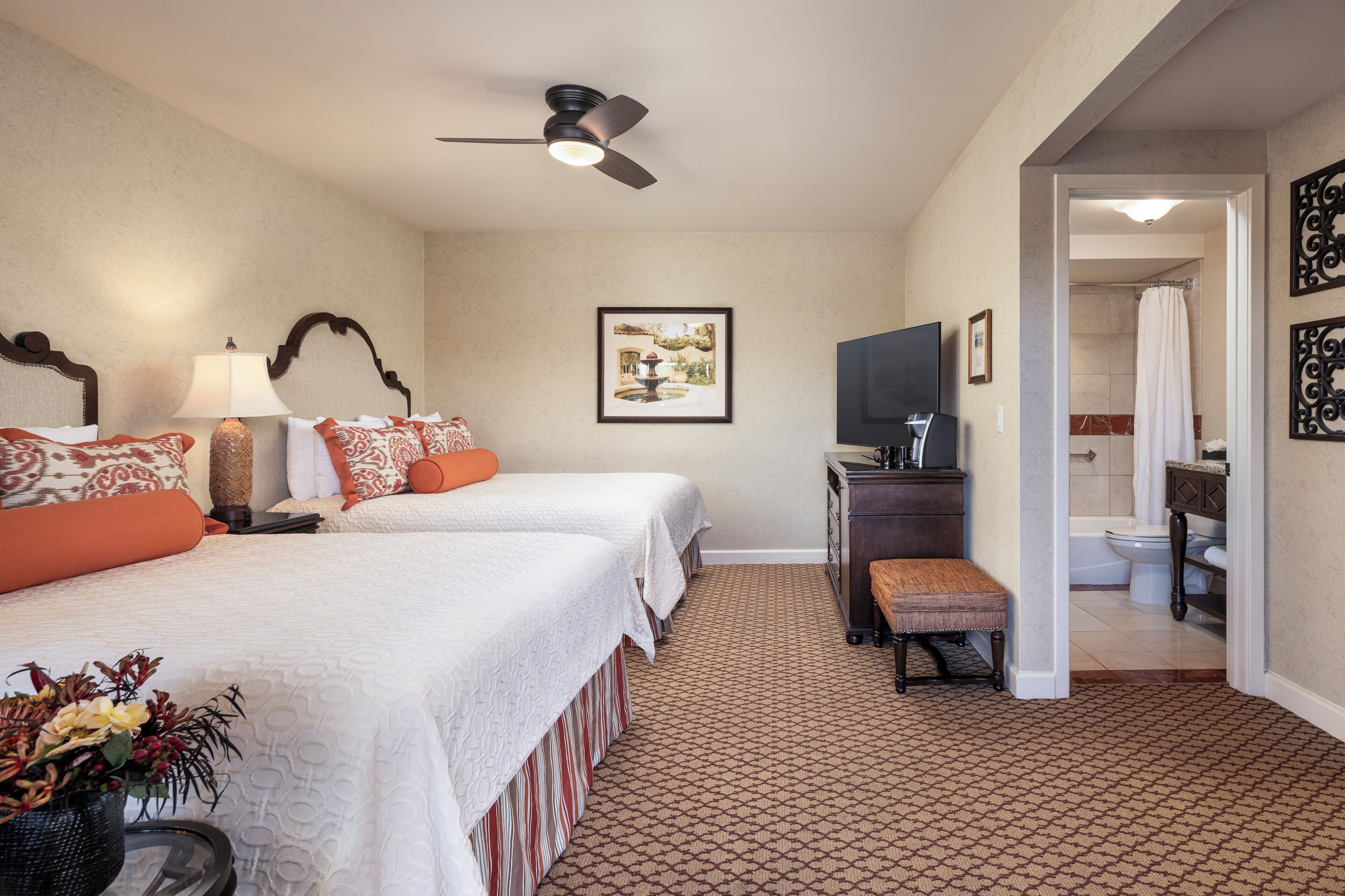 Quarto de hotel com duas camas, uma escrivaninha, televisão e banheiro visível por uma porta aberta, além de um tapete estampado.