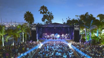 A concert stage with a large audience seated in an outdoor amphitheater surrounded by palm trees.