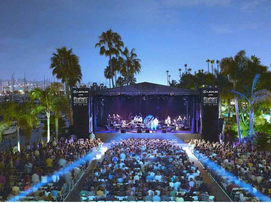 A concert stage with a large audience seated in an outdoor amphitheater surrounded by palm trees.