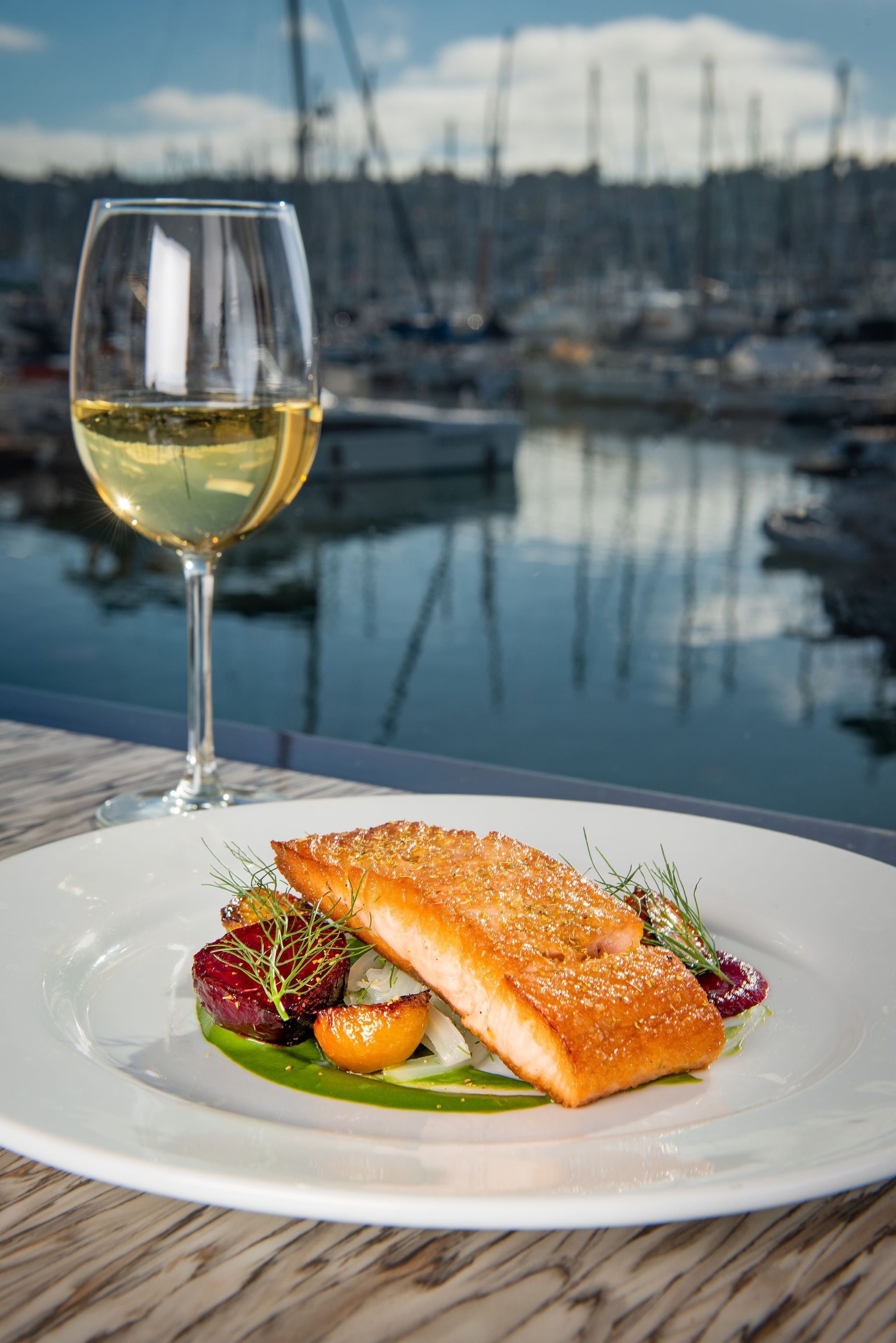 A plate of food with a glass of white wine on a table with a marina view in the background.