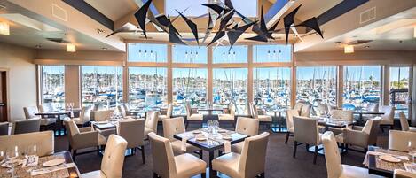 A restaurant with a view of a marina, featuring high ceilings, modern lighting, and a central chandelier.