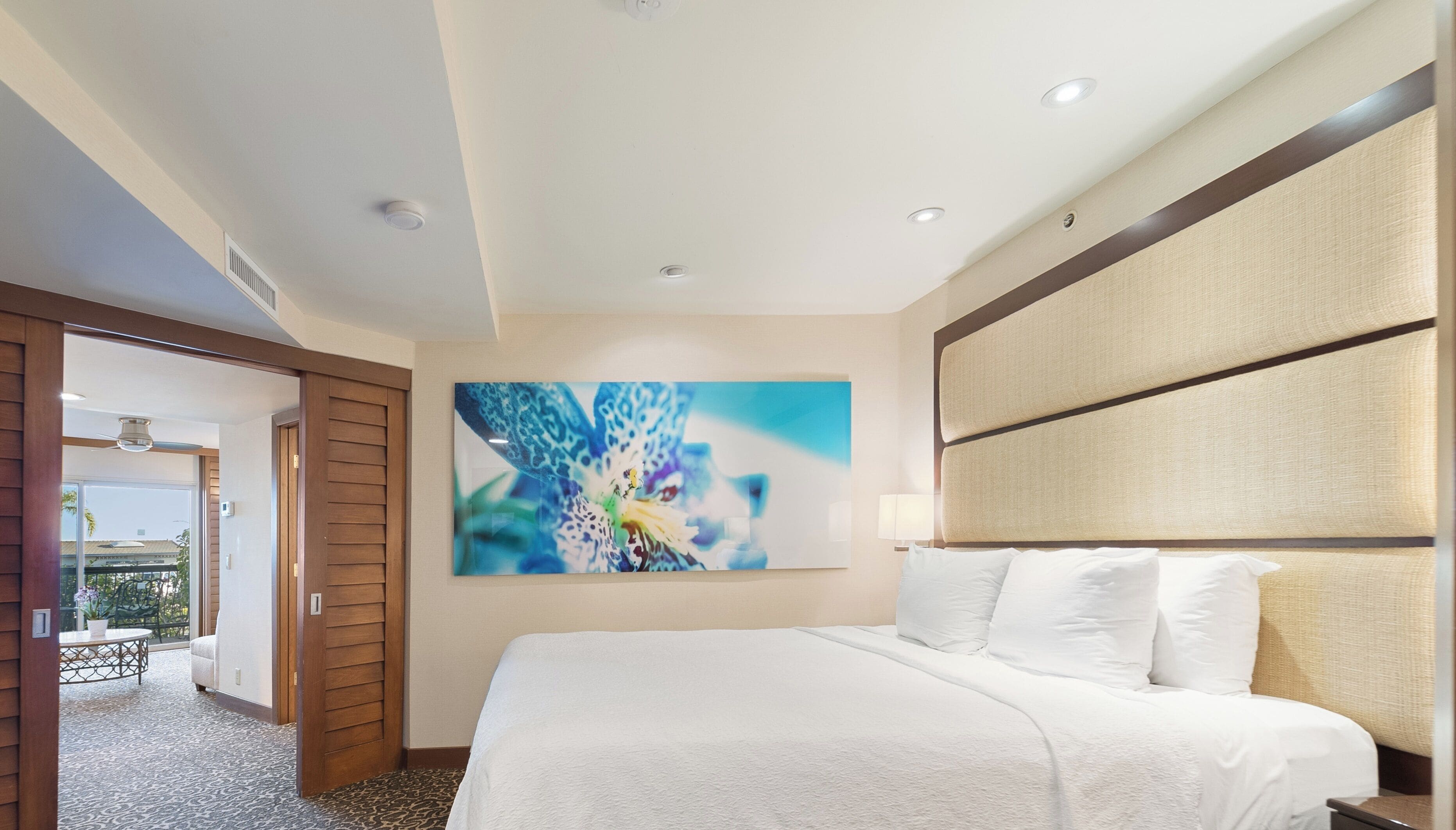 Une chambre d’hôtel avec un grand lit, un ventilateur de plafond, un tableau au mur et une vue sur l’extérieur par une porte.
