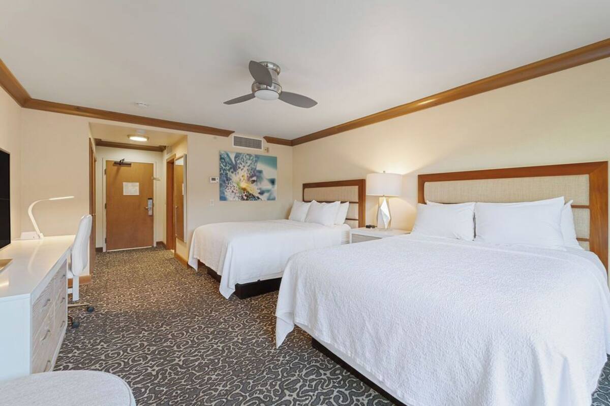 Une chambre d’hôtel avec deux lits, un bureau, une chaise, un ventilateur de plafond et un tableau au mur.