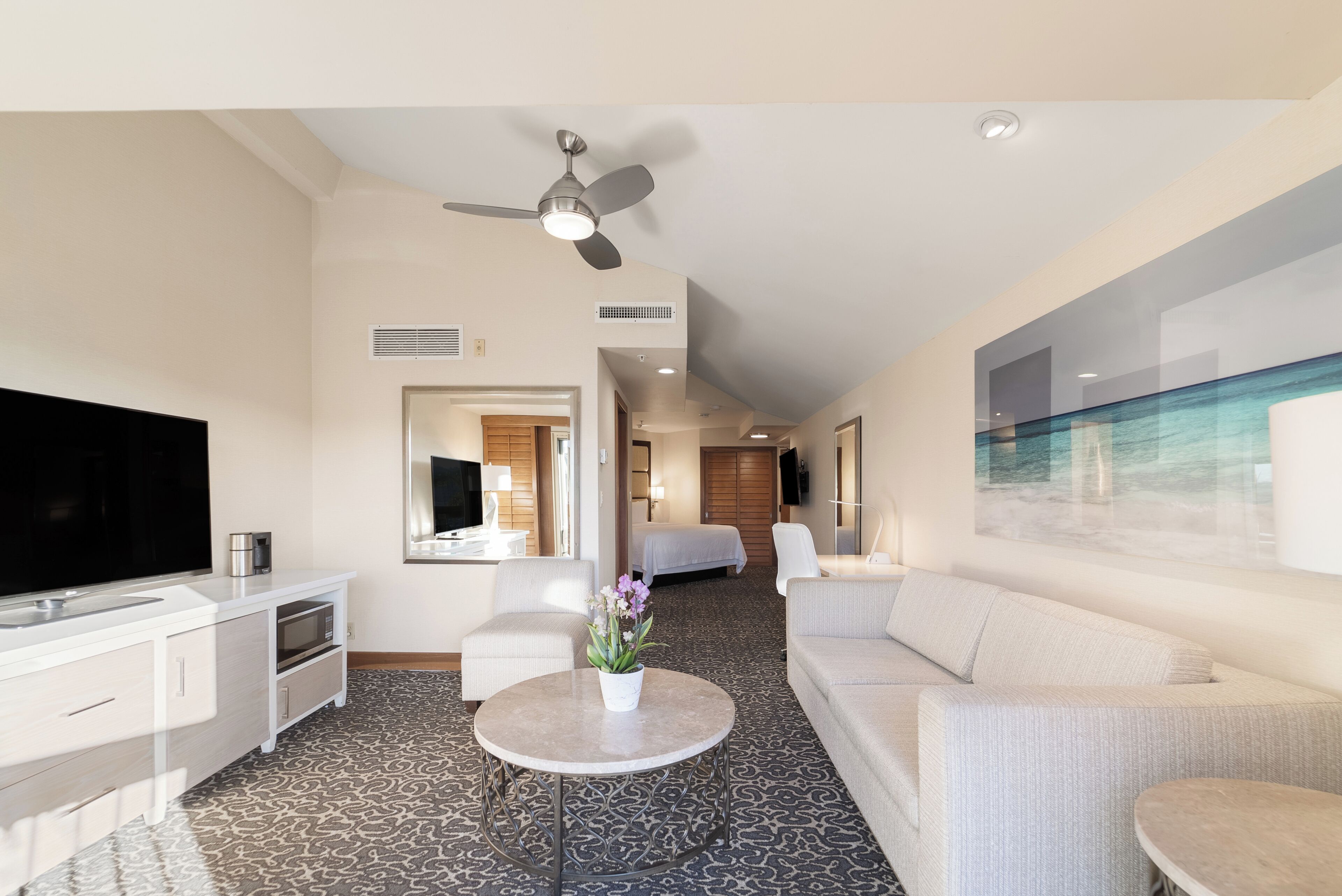 A hotel room with a flat-screen TV, a ceiling fan, a bed, a sofa, a coffee table, and a painting on the wall.