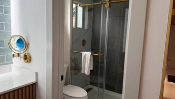 Une salle de bain comprenant des toilettes, une douche en verre avec un pommeau doré et un miroir fixé au mur, doté d’un cadre doré.