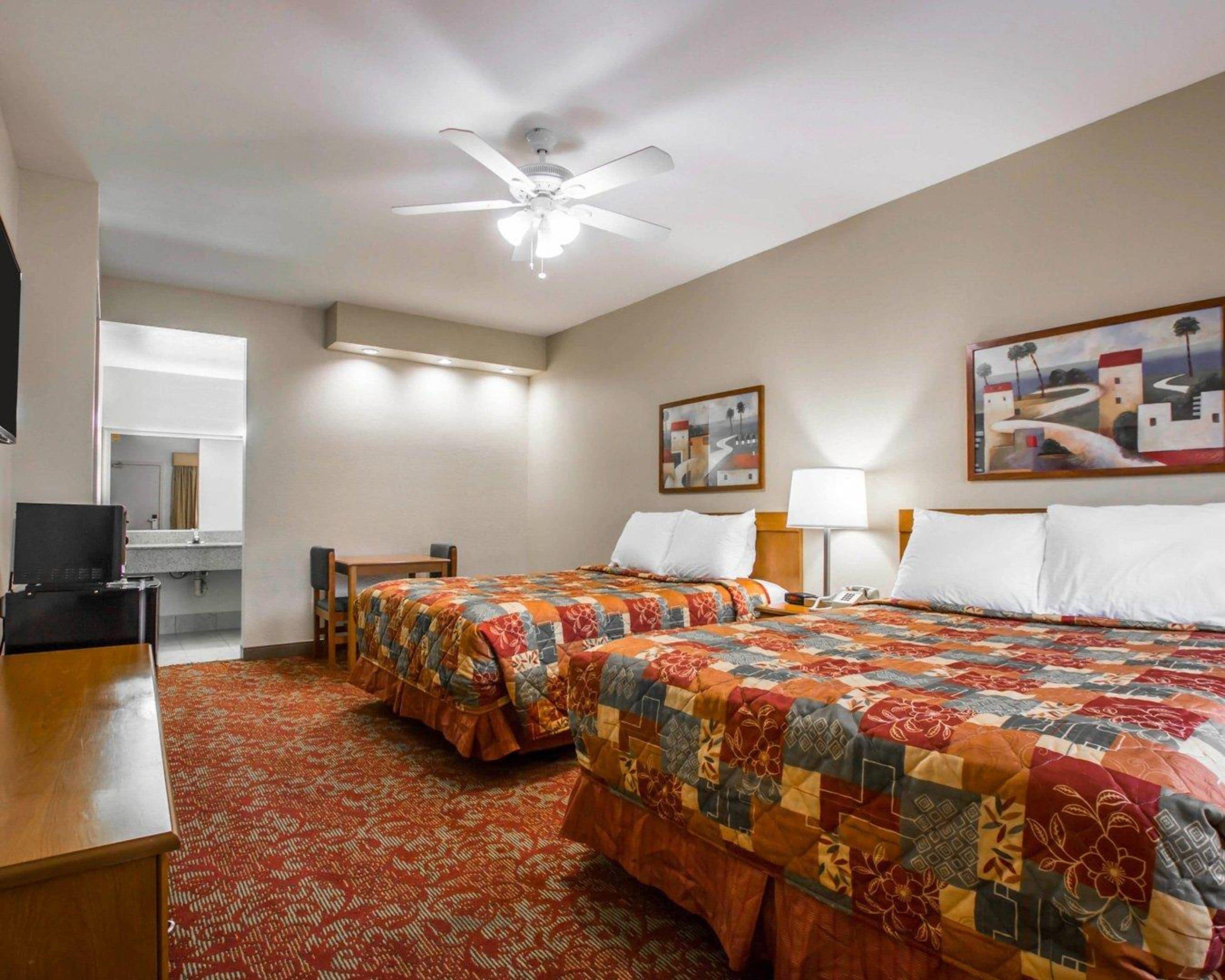 A hotel room with two beds, a desk, a chair, a ceiling fan, and artwork on the walls.