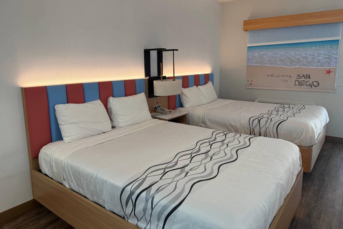 Habitación de hotel con dos camas, una mesita de noche con lámpara y un mural en la pared que da la bienvenida a San Diego.