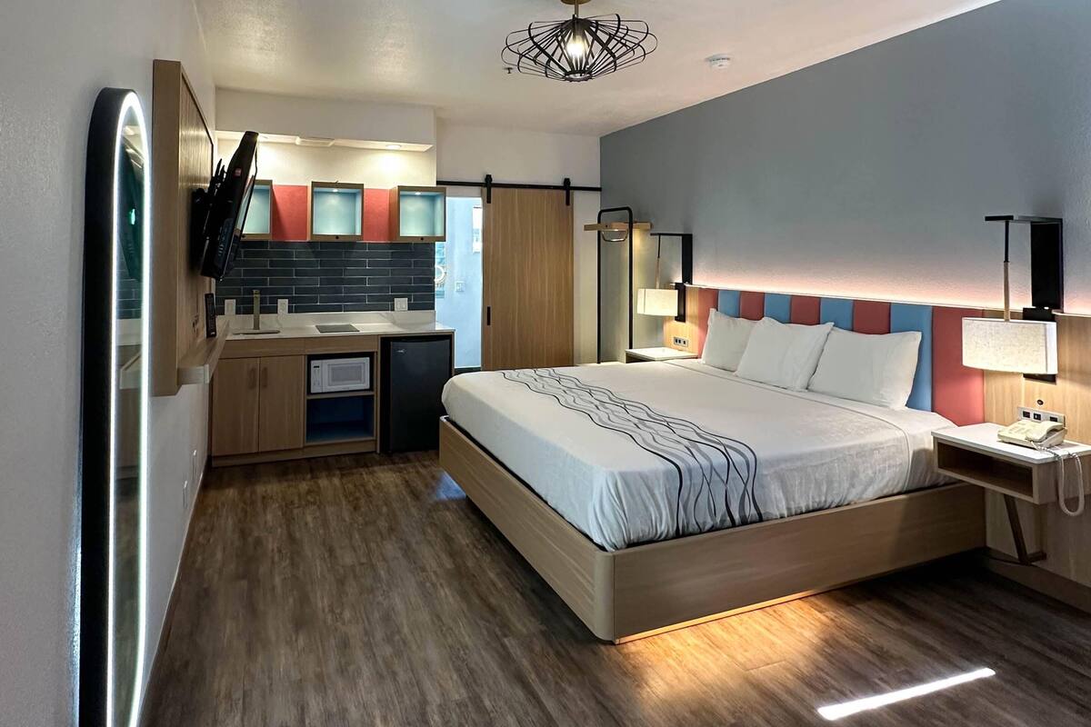 Una habitación de hotel moderna con cama, mesita de noche, lámpara y un pequeño escritorio con espejo y televisión.