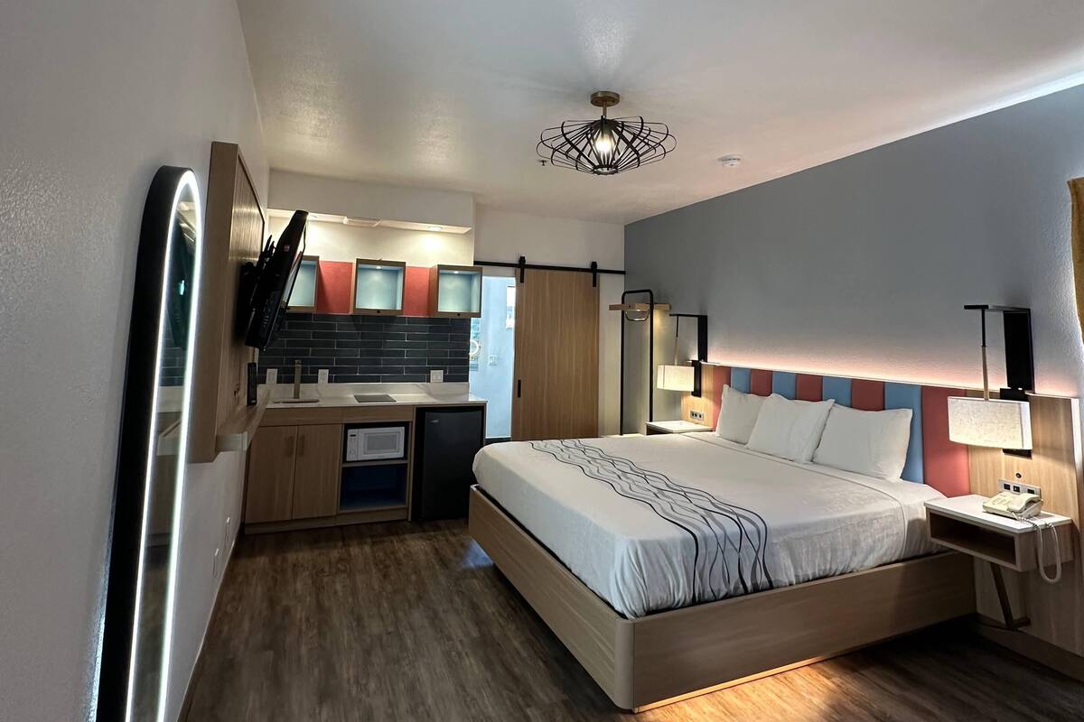 Una habitación de hotel compacta con cama, mesita de noche, lámpara y un rincón con kitchenette.
