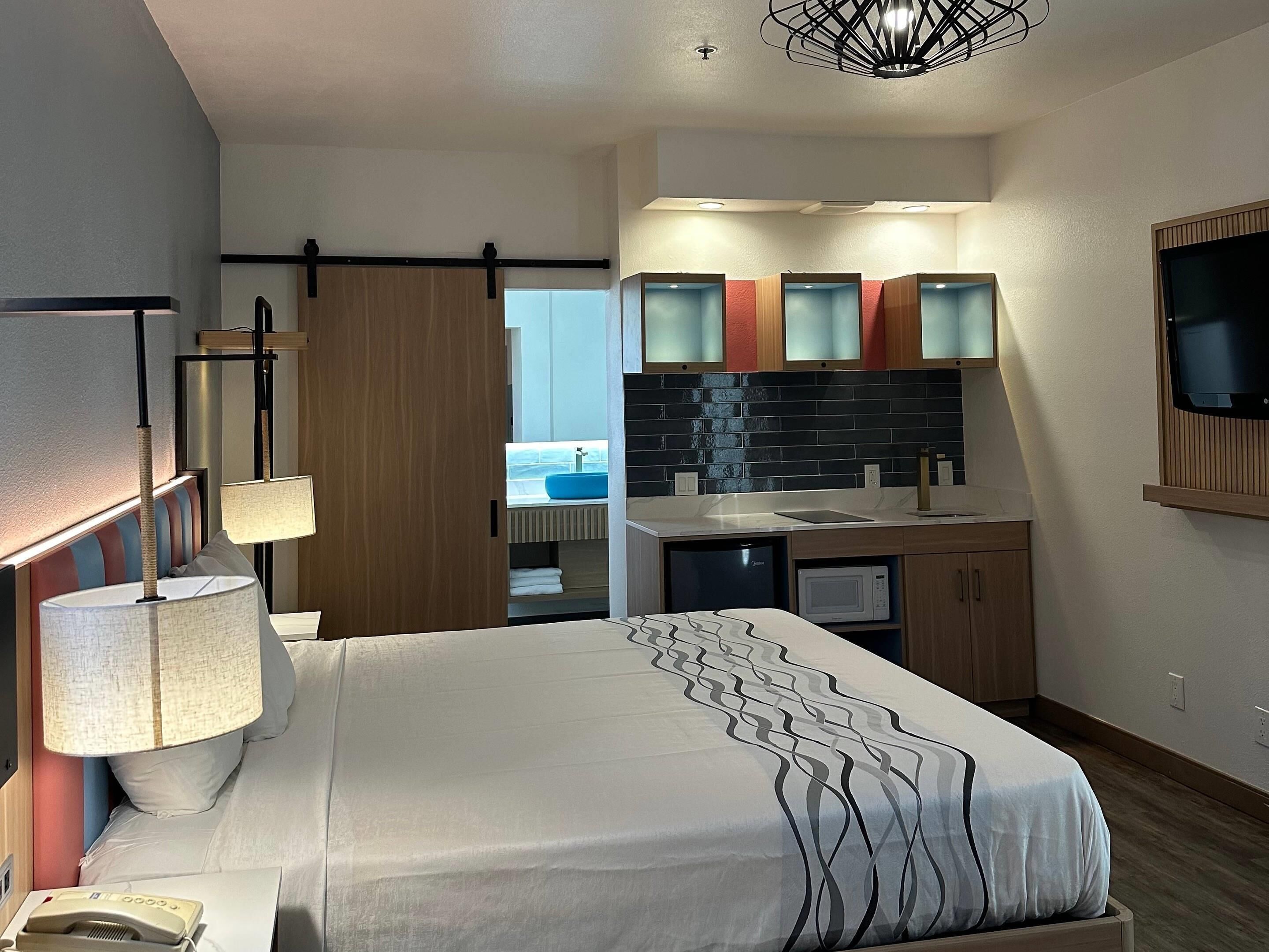 Une chambre d’hôtel moderne équipée d’un lit, d’une lampe de chevet, d’un bureau avec un ordinateur, d’un four à micro-ondes, d’un lavabo et d’un téléviseur fixé au mur.