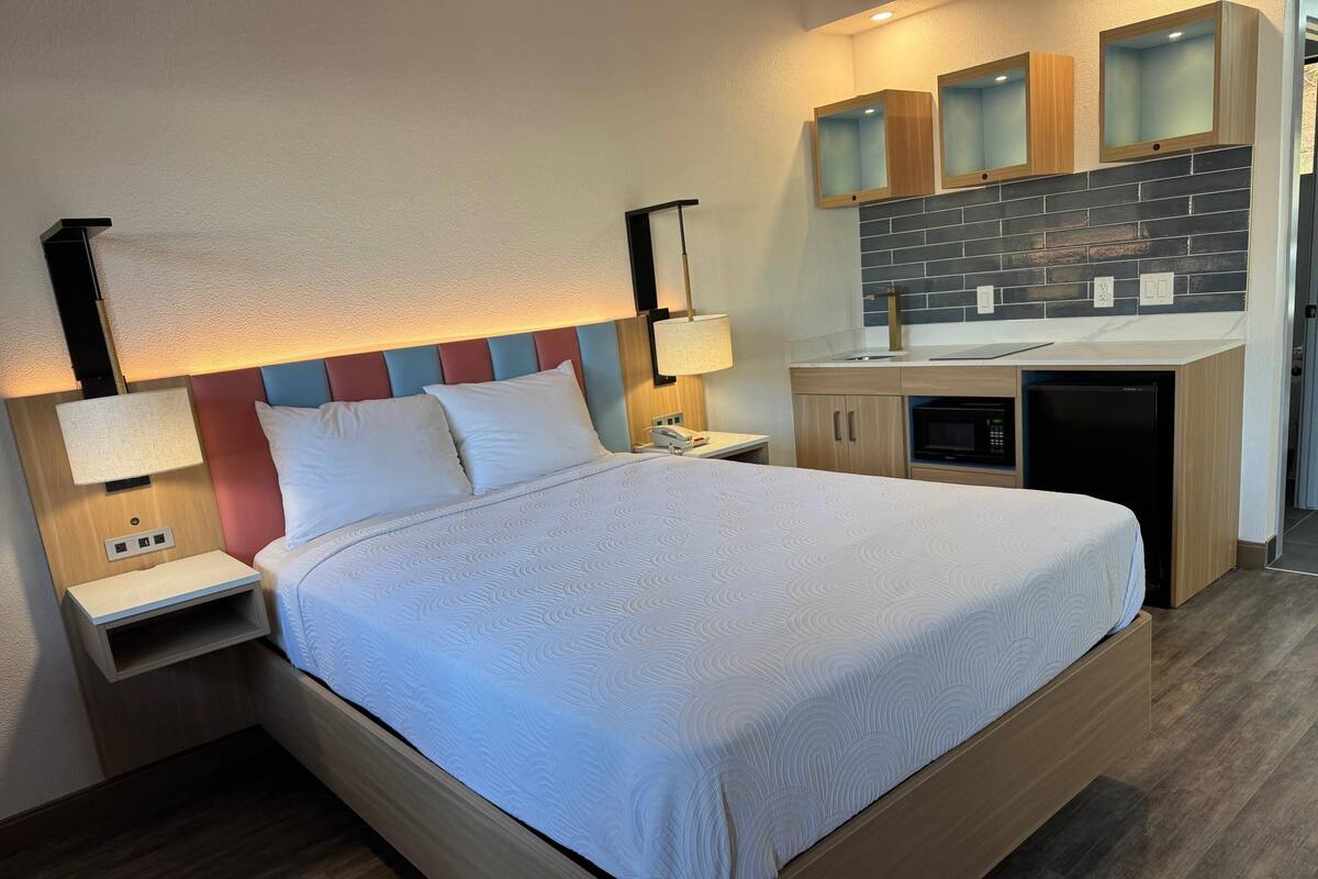 Una habitación de hotel moderna con una cama grande, mesitas de noche, un escritorio integrado, una pequeña cocina y un baño.