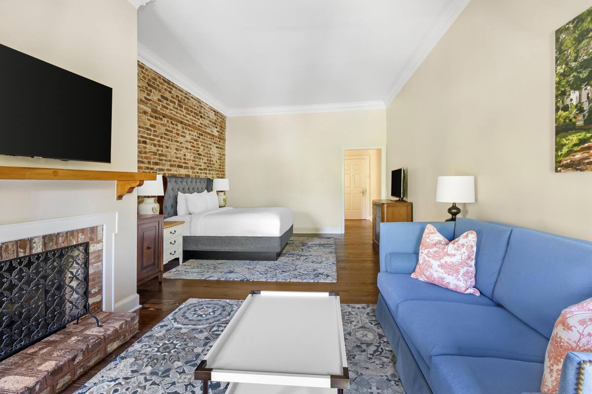 A hotel room with a bed, a blue sofa, a flat-screen TV, a fireplace, and a brick wall.