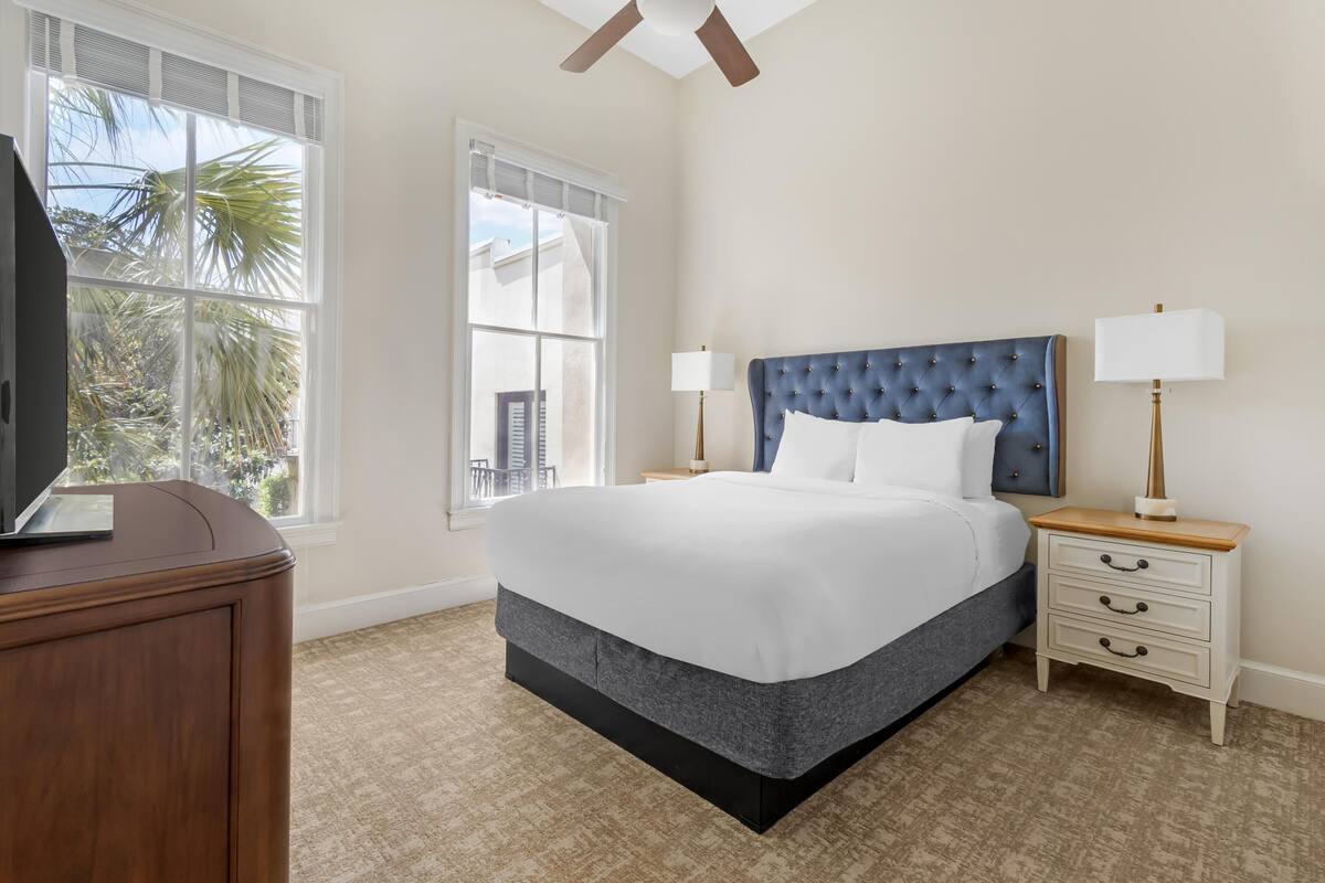 A bedroom with a large bed, two bedside tables with lamps, a TV on a wooden cabinet, and a window with a view of palm trees.