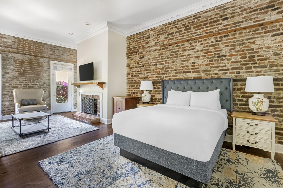 A bedroom with a brick wall, a bed, a chair, a nightstand, and a fireplace.