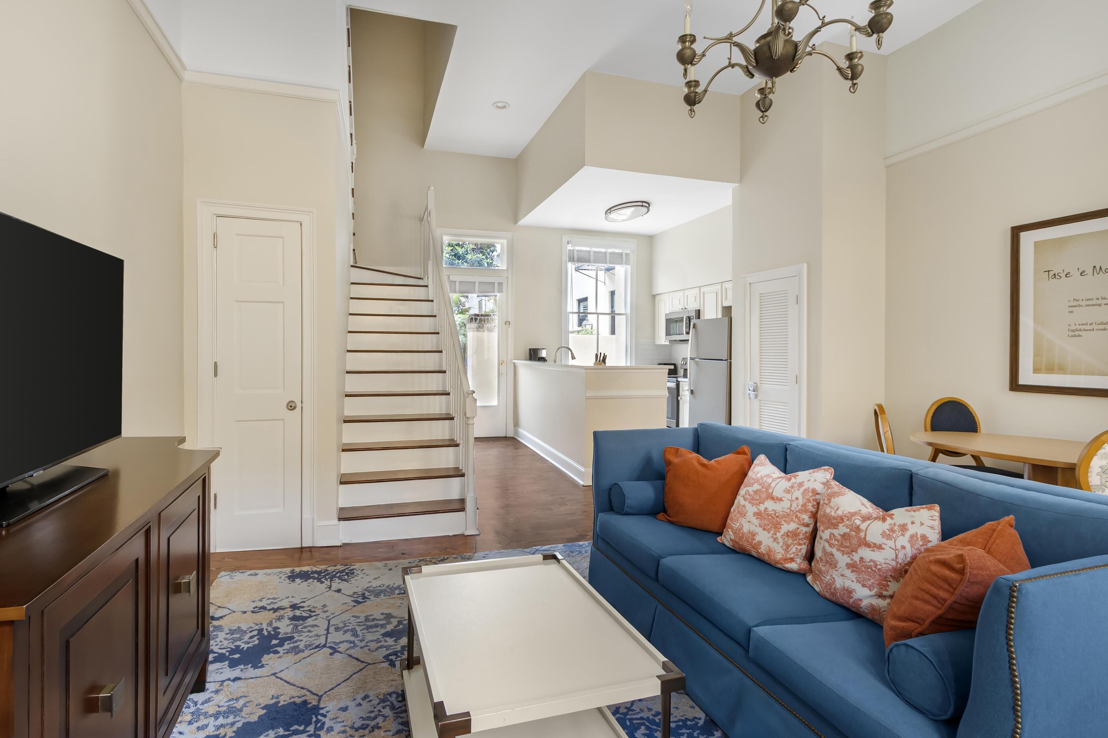 A living room with a blue sofa, a flat-screen TV, a staircase, and a kitchen in the background.