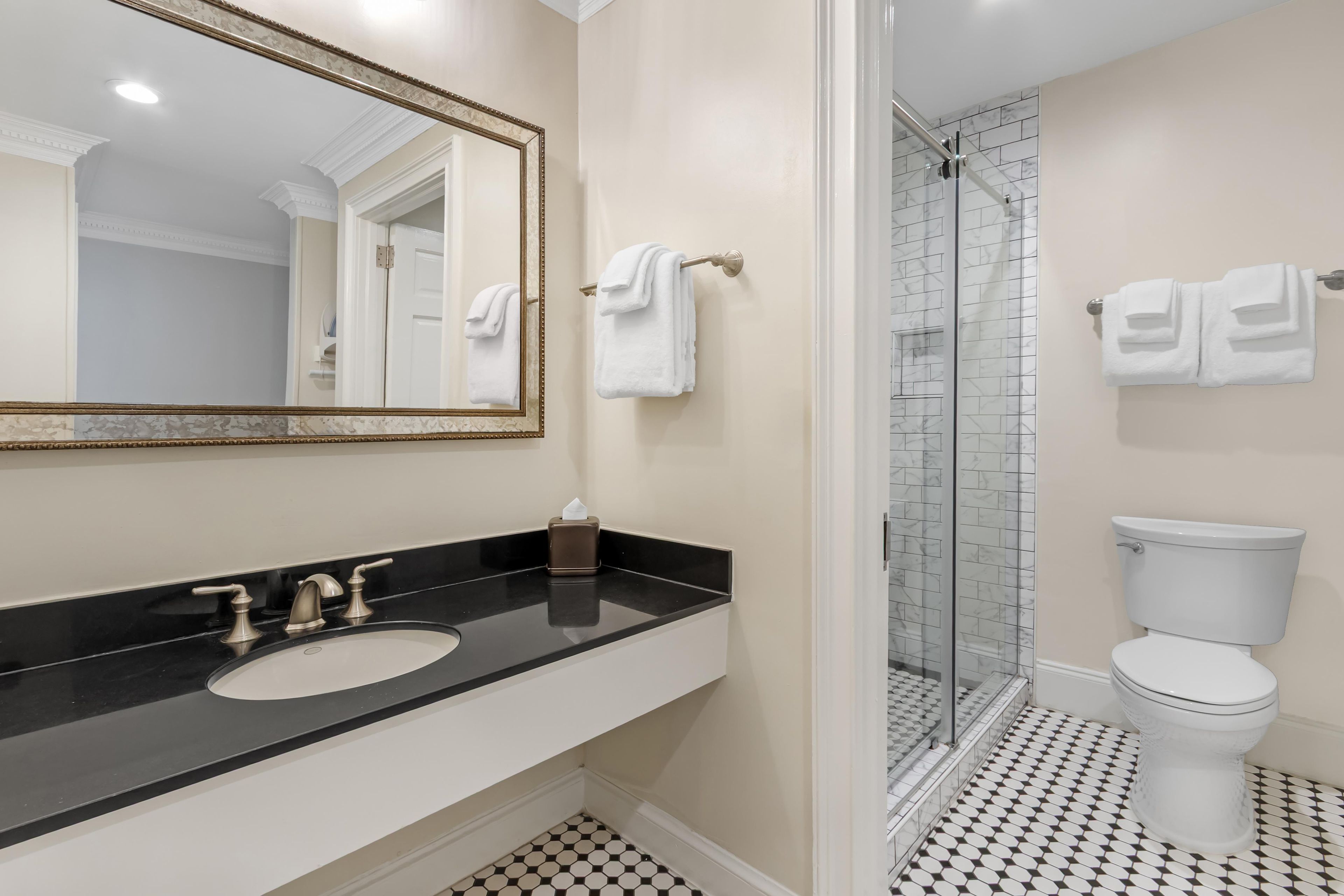 Een badkamer met een zwart aanrechtblad, een wastafel, een spiegel met gouden lijst, een wit toilet en een douchecabine met glazen wanden.