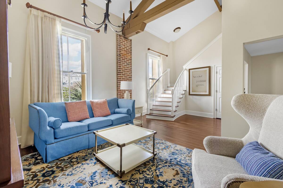 A living room with a blue sofa, a white coffee table, a patterned rug, a brick fireplace, and a staircase.